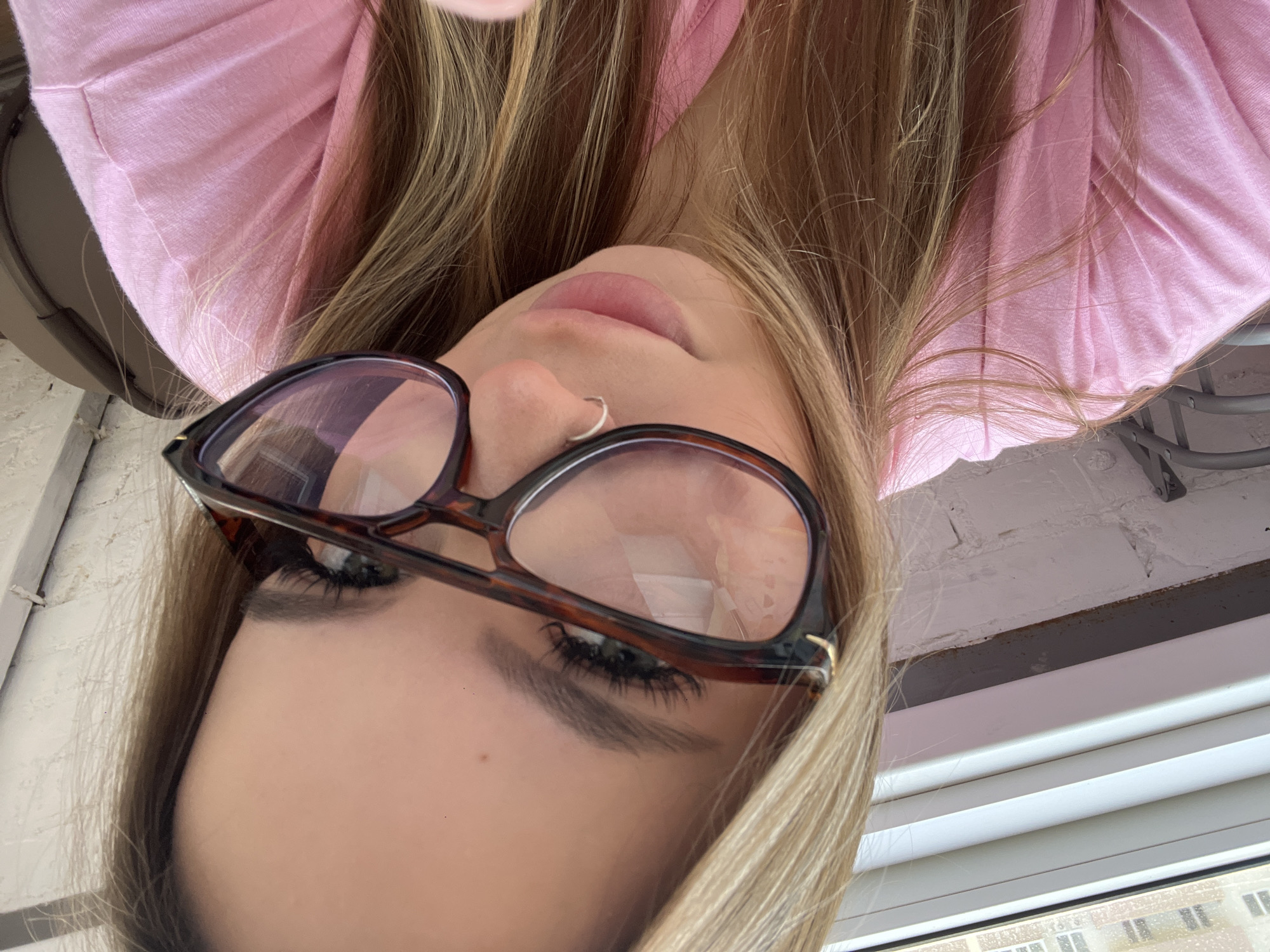A person, oriented upside down, is wearing eyeglasses with tortoise-shell frames and a silver nose ring. They are wearing a light pink top. The subject's hair is blonde with darker roots. Visible behind the person is a window and the exterior of a building, suggesting an interior setting. The location is Prigorodny, Russia.