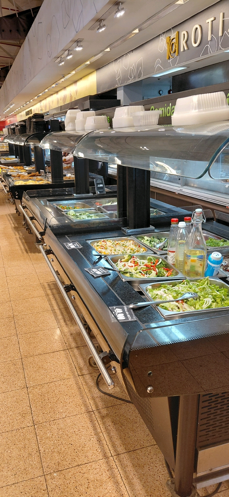 The image shows a self-service food station, likely in a supermarket or restaurant, with various salads and other prepared foods displayed in stainless steel trays. The food station has a black countertop and is illuminated by overhead lights. There are clear plastic sneeze guards over the food. In the background, there is a sign that appears to say "ROTI" and possibly other text in Spanish. The flooring is tiled with a speckled brown and beige pattern. The overall environment suggests a commercial setting for food service. The sign mentioning "Comida" (food) and a price in "4.990" (likely Paraguayan Guaraní) suggests this location is in Encarnacion, Paraguay. There are no visible people in the image.
