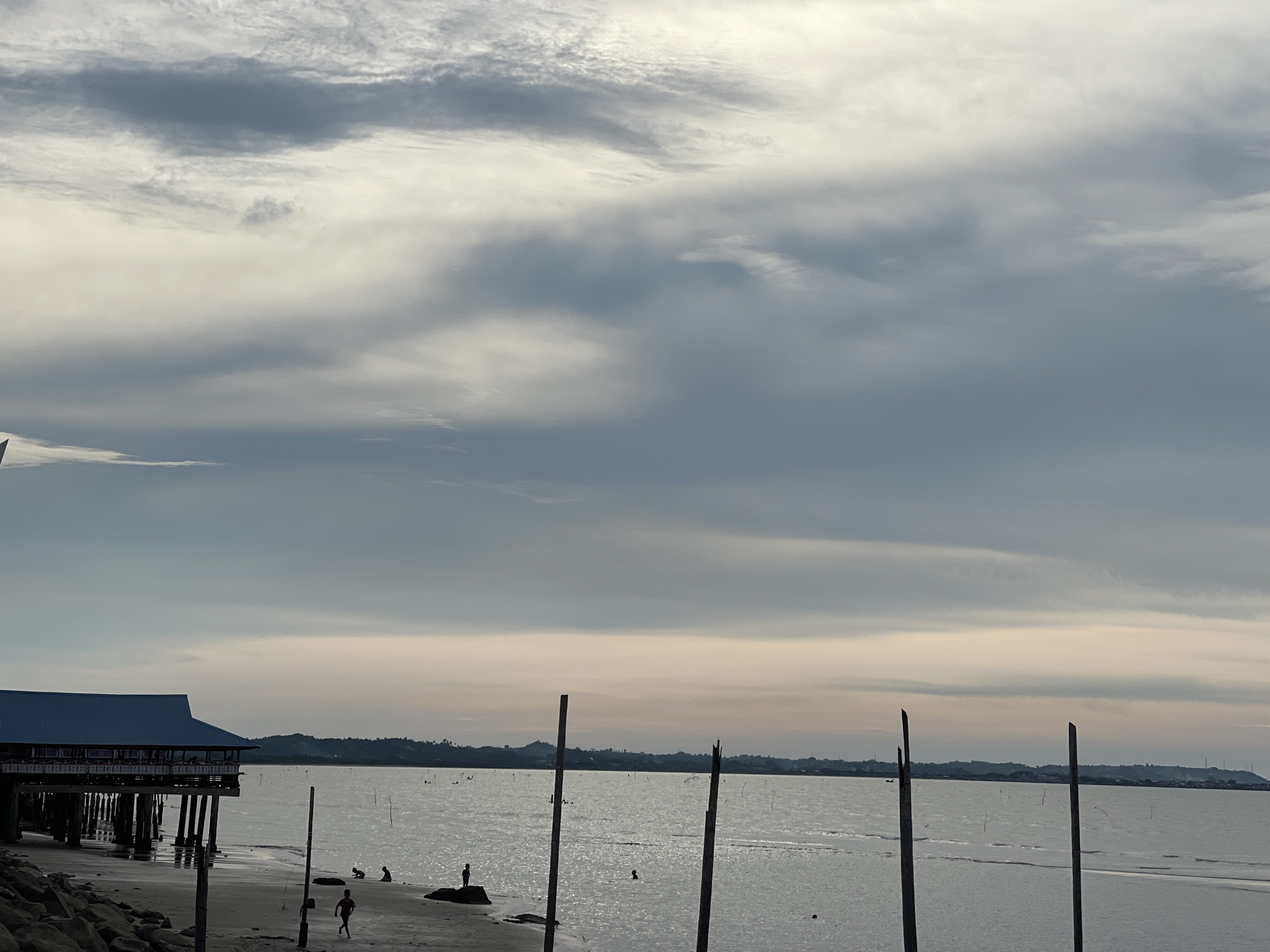 The image depicts a beach scene in Lancok, Indonesia. The sky is overcast with layered clouds. A dark blue-roofed building on stilts extends over the water from the shore on the left side of the frame. The beach in the foreground is composed of sand and rocks. Several figures, appearing to be children, are scattered across the beach and in the shallow water. Two wooden poles stand upright in the sand near the water's edge. The ocean stretches to the horizon, where a distant, hilly coastline is visible under the diffused light.