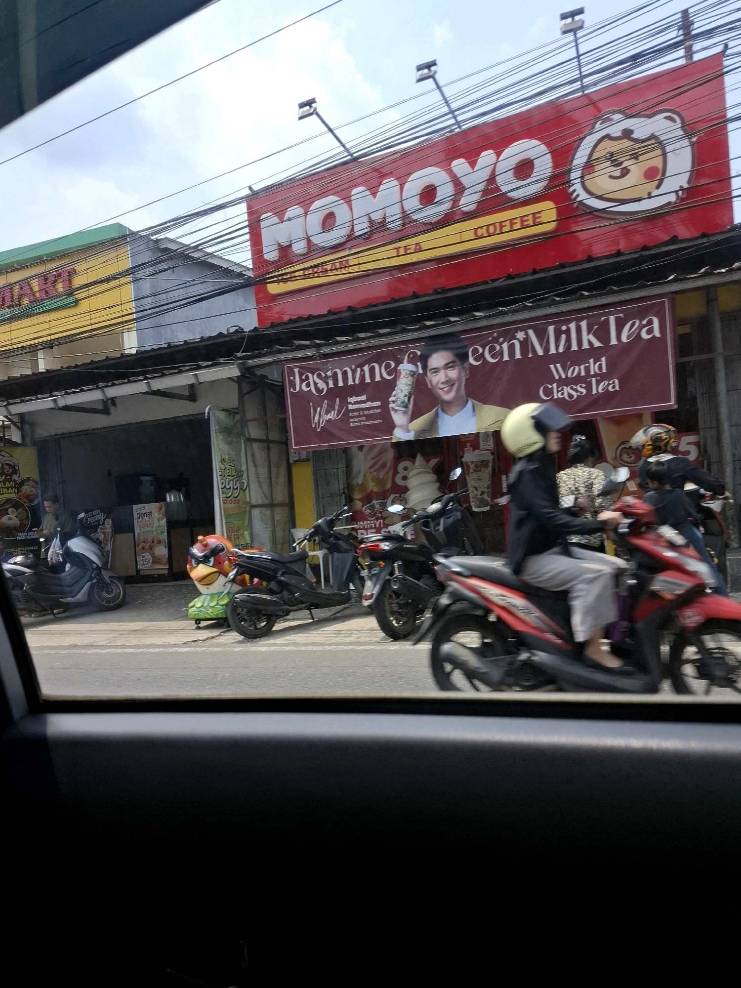 This is a daytime, eye-level, medium shot of a street scene captured through a car window. In the foreground, the dark interior of the car is visible, with the car's side panel dominating the lower portion of the frame. The middle ground depicts a busy street in Sukabumi, Indonesia, characterized by multiple parked and moving motorbikes. On the right, a red motorbike with a rider wearing a yellow helmet and a black jacket is in motion. To the left, a silver motorbike is parked, and another black motorbike is also visible. Buildings line the street, featuring various signs. A prominent red billboard at the top displays the logo "MOMOYO" and the words "COFFEE" and "TEA" in white and yellow text, accompanied by a cartoon character. Below this, a maroon banner advertises "Jasmine Green* Milk Tea" with an image of a man holding a drink. In the background, shops and awnings are visible, along with electrical wires crisscrossing overhead. The sky is partially cloudy.