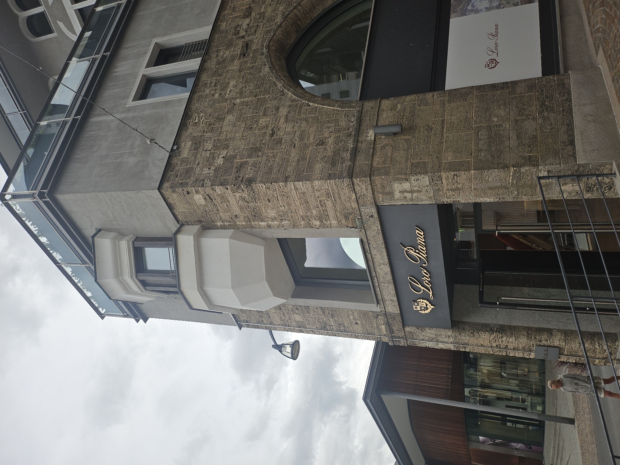 This is a vertical, medium shot of a building's facade in St. Moritz, Switzerland. The building features a mix of architectural styles. The upper portion has a modern, glass-enclosed section that angles upwards, contrasting with the rough, textured stone facade below. A prominent circular window is set into the stone. A sign with the text "Loro Piana" in gold lettering is mounted on a dark surface above a shop entrance. A large poster or advertisement featuring "Loro Piana" with a logo is visible in an upper window. The scene appears to be outdoors, with a cloudy sky suggesting overcast weather. A street lamp is visible attached to the building. In the lower right of the frame, a person is walking on a paved area in front of the shops. The overall impression is a street view of a commercial building in an urban or semi-urban setting.