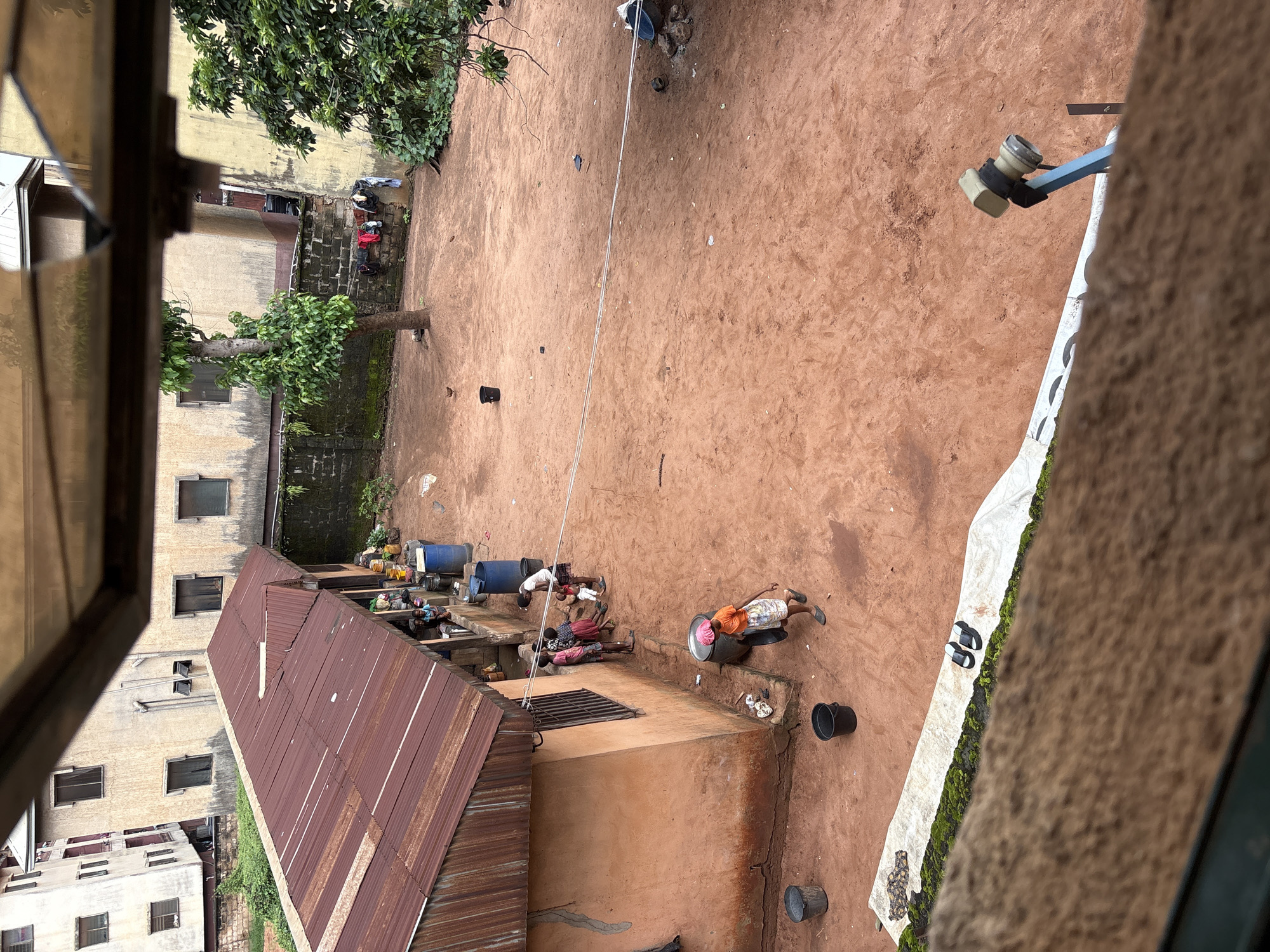 This is a medium-shot, high-angle photograph taken outdoors in what appears to be a courtyard or open space in Onitsha, Nigeria. The ground is composed of reddish-brown earth.
In the foreground, a white tarp is laid out, with green moss or vegetation growing along its edge. A pair of black flip-flops rests on the tarp.
Dominating the middle ground is a building with an orange-colored stucco exterior, featuring a window with bars. To the left of this building, a section of a long, low structure with a rusty, corrugated metal roof extends outwards. Several individuals, appearing to be adults and children, are visible near this structure, some standing and interacting, others sitting. A large blue barrel is situated near the structure, and other buckets and containers are scattered around.
Further back, in the background, are multi-story buildings with beige-colored exteriors and dark-framed windows. A mature tree with green foliage stands in the courtyard, and laundry hangs on a line strung between two points in the background, near a brick wall.
One person in the foreground, a woman wearing an orange top and a patterned skirt, walks away from the orange building. Another person, partially visible, appears to be sweeping or cleaning the ground.
The overall impression is a scene of daily life in a residential or semi-commercial area, characterized by its earthy ground, simple structures, and human activity.