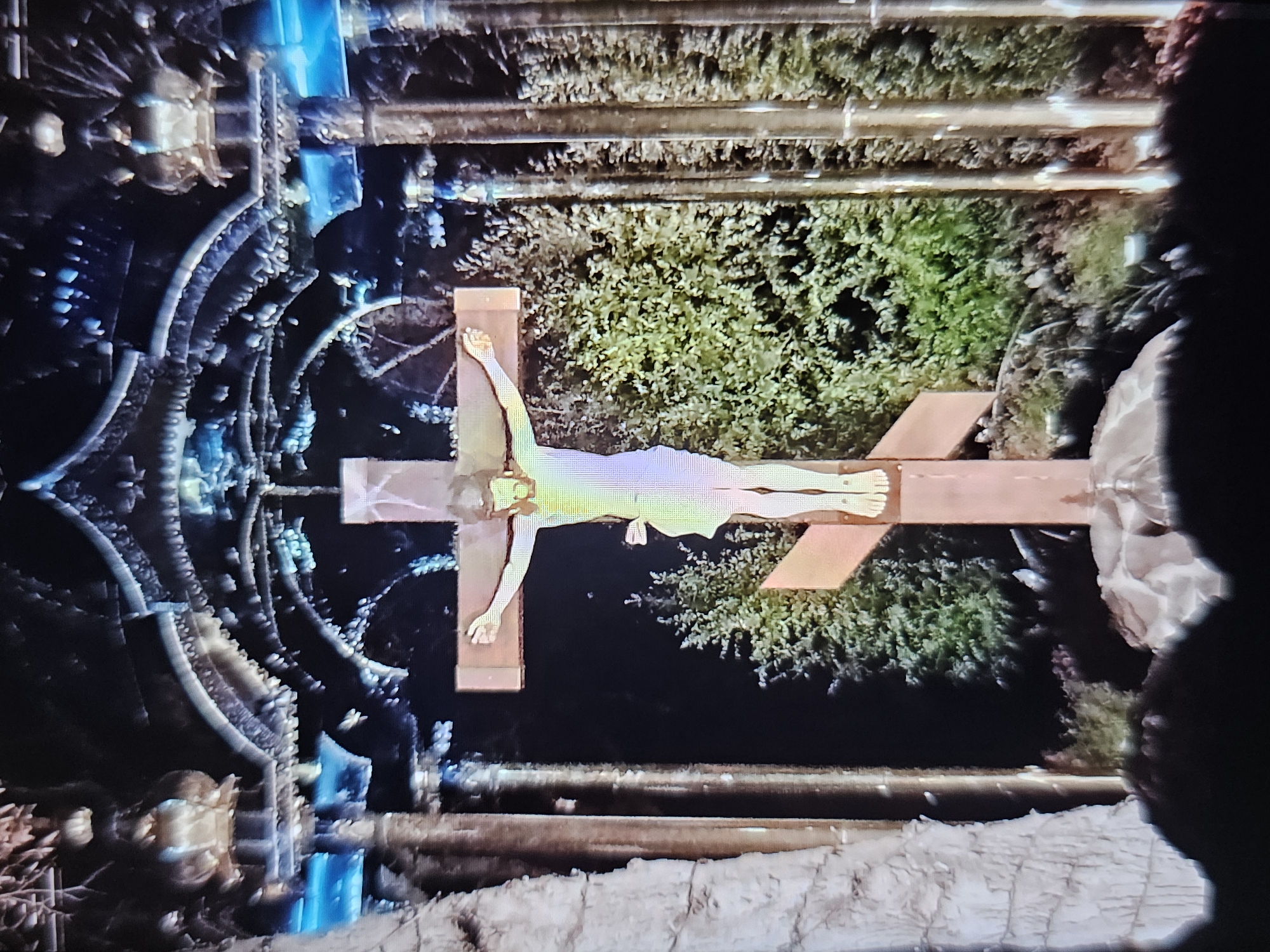 A crucifix stands in the center of the frame, featuring a figure of Jesus Christ. The crucifix is made of wood and has an additional diagonal crossbar at the bottom, characteristic of Orthodox Christian iconography. Behind the crucifix, lush green trees are visible. The scene is framed by elaborate architectural elements overhead, which appear to be ornate metalwork with blue lighting. To the left of the crucifix, a large, textured column is present, possibly made of stone or a similar material. The lighting suggests it is nighttime or a dimly lit indoor space. This image was taken at The Big Texan Steak Ranch in Amarillo, Texas, United States.