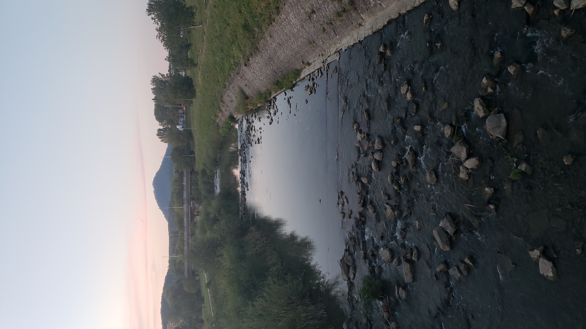 The image captures a wide-angle view of a river flowing through an urban environment, likely Prešov, Slovakia, during the late afternoon or early evening. The sky is a soft gradient of pale blue and pink, indicating the setting sun.
In the foreground, the river is characterized by a rocky bed, with numerous stones of varying sizes breaking the surface of the dark water. Small ripples and currents are visible around these rocks, suggesting a moderate flow. The riverbanks are lined with lush green vegetation, particularly dense on the left side, and a concrete embankment on the right.
Further upstream, a low bridge crosses the river, with traffic lights and streetlights visible on its approach. Beyond the bridge, a prominent conical hill dominates the horizon, characteristic of the landscape surrounding Prešov. Trees of various sizes are scattered along the banks and on the slopes of the hills, adding to the natural elements of the scene. The overall impression is a serene, yet slightly melancholic, urban landscape at dusk.