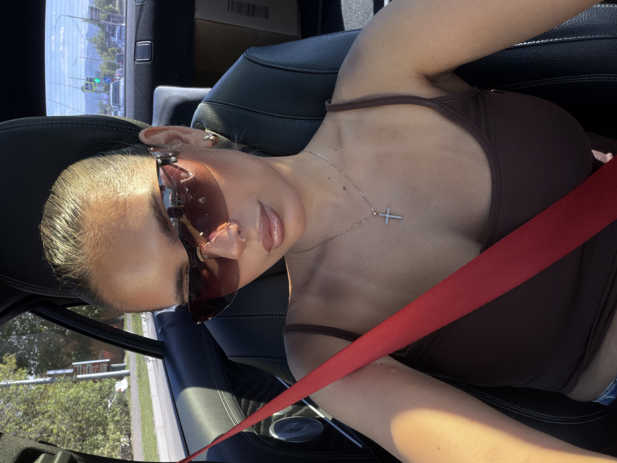 A woman is sitting in the driver's seat of a car. She is wearing a brown tank top, a silver cross necklace, and sunglasses. She has a red seatbelt buckled across her chest. The car's interior is black leather. Outside the car window, a street scene is visible. Based on visual clues, this is not Krasnoyarsk, Russia.