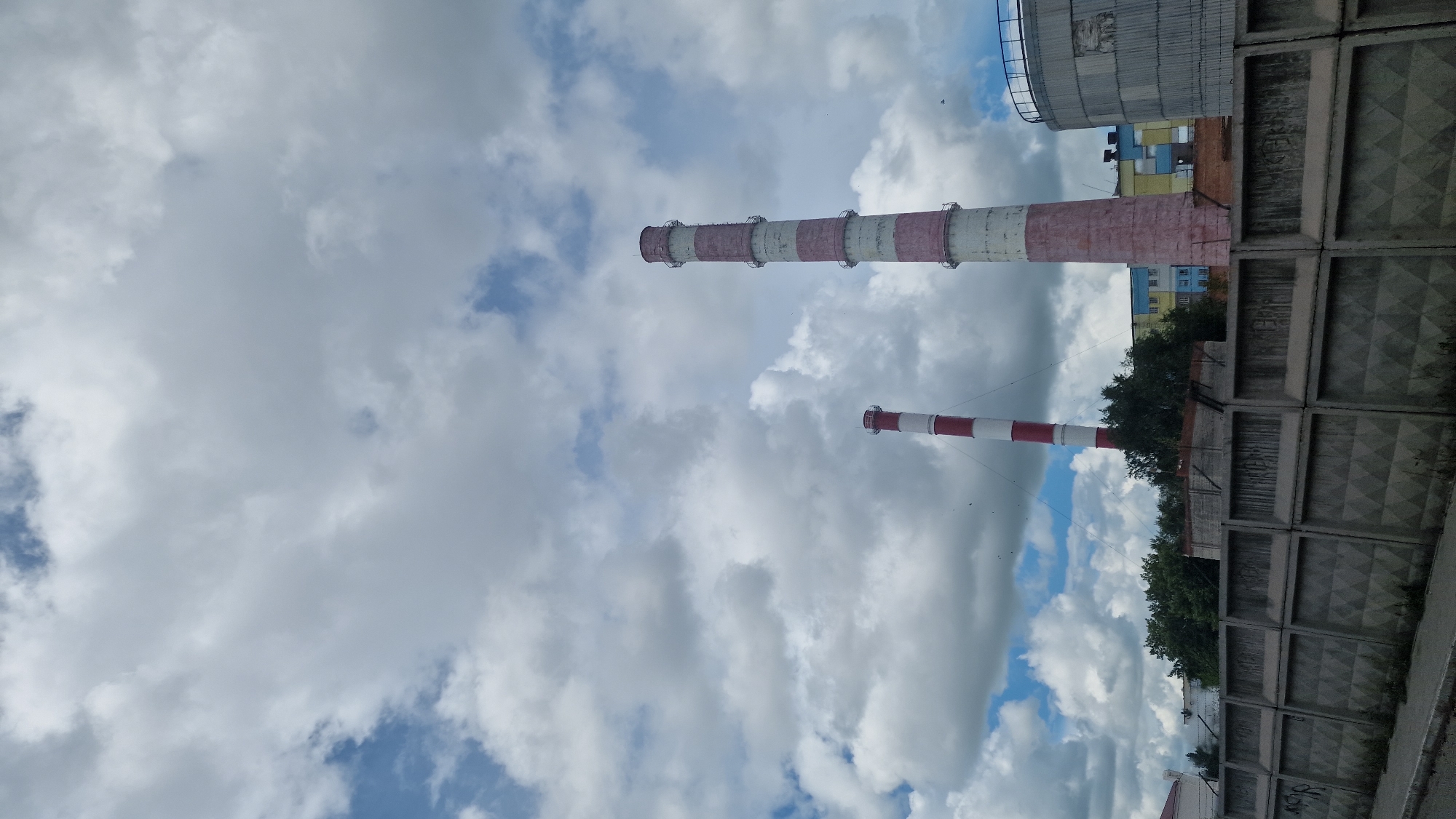 Two tall industrial smokestacks, one larger and pink with white bands, and a smaller one with red and white bands, dominate the upper portion of the frame against a cloudy sky. In the middle ground, behind a grey concrete fence with diamond patterns, is a cluster of buildings, some colorful with windows and a cylindrical structure to the right. Green trees partially obscure some of the buildings. The foreground shows a dark asphalt surface and the base of the concrete fence. The scene appears to be in an industrial area within the city of Kostanay, Kazakhstan.