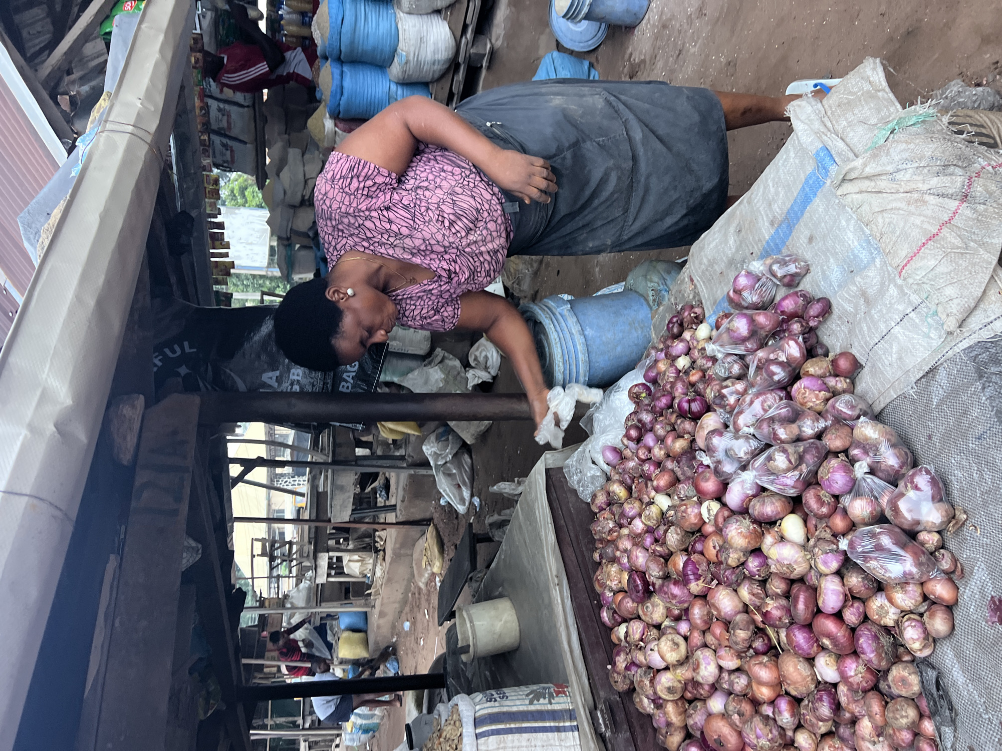 In Nkpor, Nigeria, a woman in a pink patterned blouse and a dark skirt is seen at a market stall. She is interacting with a large pile of red onions, some of which are bagged. The stall is covered by a corrugated metal roof supported by wooden beams. Various sacks, possibly containing grains or other produce, are visible in the background, along with other market vendors and stalls. The ground appears to be dirt. The woman is holding a white plastic bag in her right hand.