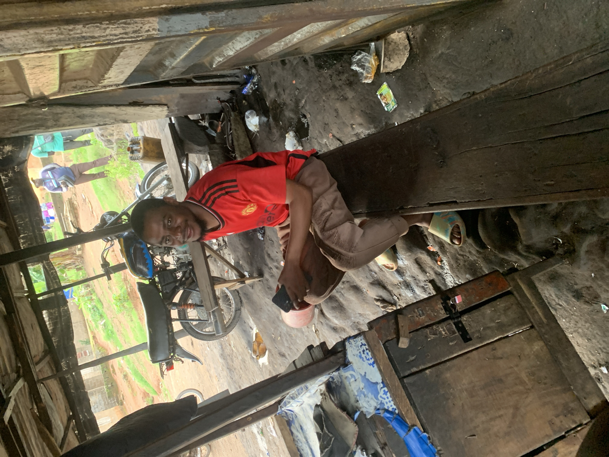 This media file depicts a scene within a rustic workshop or open-air structure, likely in Shagamu, Nigeria. The primary subject is a young man wearing a red t-shirt with black stripes on the sleeves and light brown pants. He is seated and looking towards the camera with a neutral expression. His right hand is holding a black smartphone over a small, pink bucket.
The setting appears to be an informal workspace, characterized by wooden structures, a dirt floor, and scattered debris. In the background, there are glimpses of what seems to be an outdoor environment with greenery and some buildings, suggesting a rural or semi-urban location. Several motorcycles are visible, indicating that the workshop might be involved in motorcycle repair or sales. One motorcycle is particularly prominent, with its front wheel and part of the frame visible.
Other individuals are partially visible in the background, including someone wearing a blue jacket and another in a green top, further suggesting a communal or busy setting. The overall impression is one of everyday life and work in a developing area. The lighting suggests it could be daytime, with natural light filtering into the structure. There is no discernible text visible in the media file. The scene conveys a sense of authenticity and a snapshot of daily activities within this particular Nigerian context.