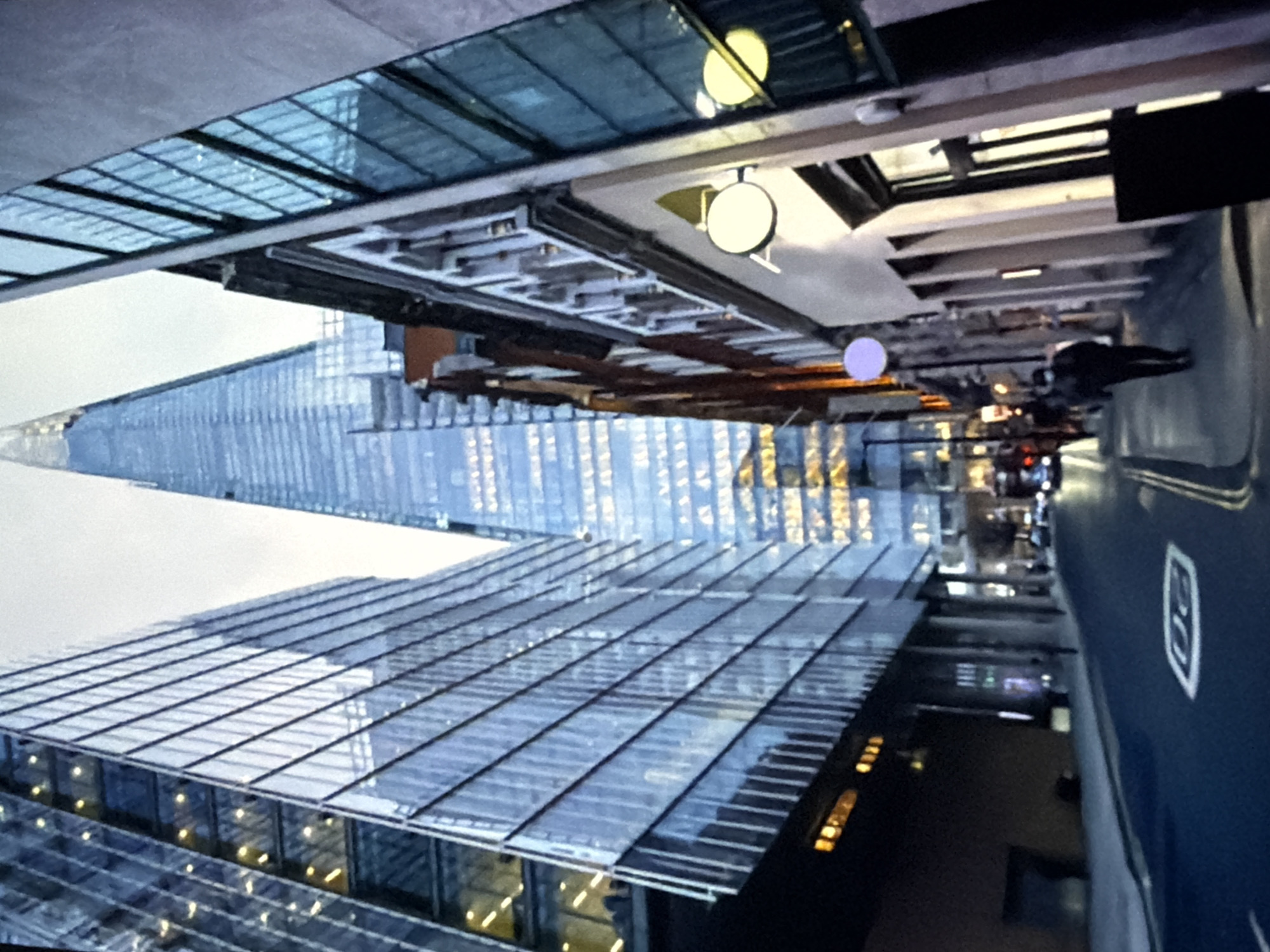 The image depicts an urban street scene at dusk, looking down a thoroughfare flanked by buildings of varying architectural styles. In the background, prominently centered, stands The Shard, a tall, tapered skyscraper with a glass facade reflecting a cool blue hue and numerous illuminated interior spaces. To the left, a large, multi-story modern building with a reflective glass exterior borders the street, displaying warm yellow interior lighting through its windows. On the right, a row of older, multi-story buildings with more traditional window designs and light-colored facades extends along the pavement.
The asphalt road in the foreground contains a white "20" speed limit marking and double yellow lines on the right side. Pavements run alongside both sides of the road. On the right pavement, several circular, illuminated signs are affixed to the buildings; one is purple, and two are white/yellow. A black A-frame sign stands on the pavement near the right edge. Further down the street, multiple vehicles are visible, indicated by their illuminated headlights and taillights. One pedestrian, wearing dark clothing and potentially a face mask, walks away from the viewer on the right pavement. Other blurred figures are discernible in the distance on the pavements. The sky visible between the buildings at the top is overcast and light grey. The scene is located in London, United Kingdom, evidenced by the presence of The Shard.