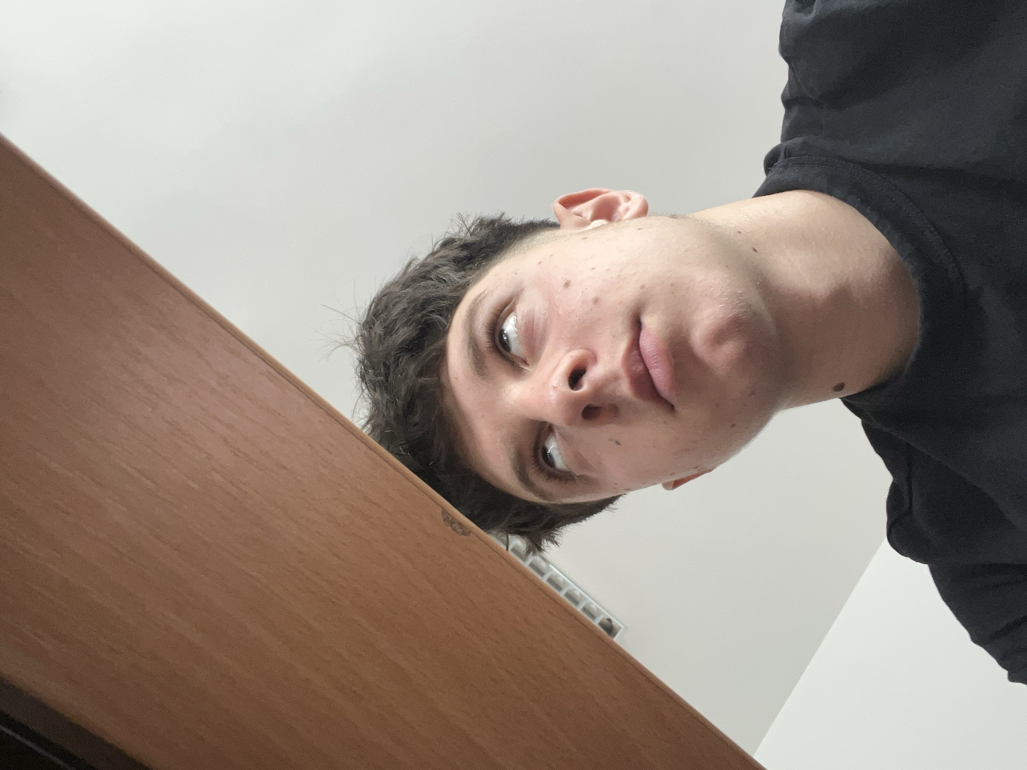 A young man with short, dark, messy hair looks upwards with his eyes rolled back, appearing contemplative or surprised. He has fair skin with some blemishes and is wearing a black t-shirt. The image is taken from a low angle, with a portion of a light brown wooden surface dominating the upper left of the frame. The background consists of a plain white wall and ceiling, with a visible vent. The setting does not provide any specific clues to its location within Zlatograd, Bulgaria.