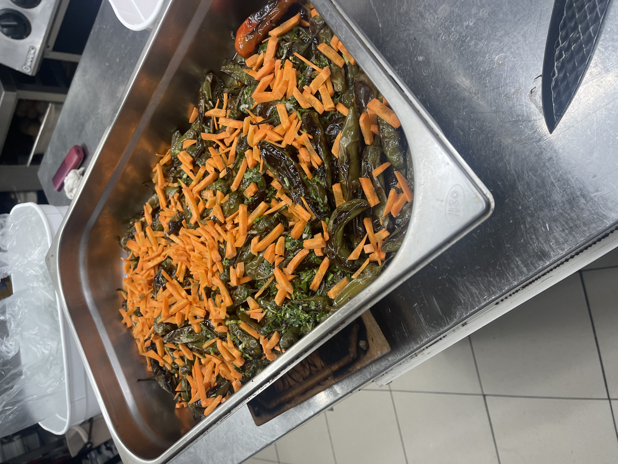 A large, rectangular stainless steel food pan, likely a hotel pan, is prominently displayed, filled with a cooked dish. The dish consists primarily of dark green, roasted or cooked long peppers, mixed with finely chopped green herbs, possibly cilantro or parsley. A generous topping of bright orange, julienned carrots is scattered across the entire surface of the food. In the upper left corner of the pan, a single roasted red pepper or tomato is visible.
The pan sits on a metallic, possibly stainless steel, counter or work surface, which shows signs of wear and scratches. To the right of the pan, the black handle and textured blade of a kitchen knife rest on the counter. In the blurred background on the left, several white plastic containers are visible, along with a small pink object and a white cloth on the counter. A partial view of a control panel with the brand "WARING" printed on it can be seen in the far upper left. Underneath the right side of the pan, a dark wooden cutting board is partially visible. The rim of the pan has the text "OZTI" embossed on it. The floor in the background features light-colored square tiles.
The setting appears to be a commercial kitchen or a food preparation area. No people are visible, and there is no specific activity taking place, suggesting the food is prepped and ready for serving or further use. The lighting indicates an indoor environment.
