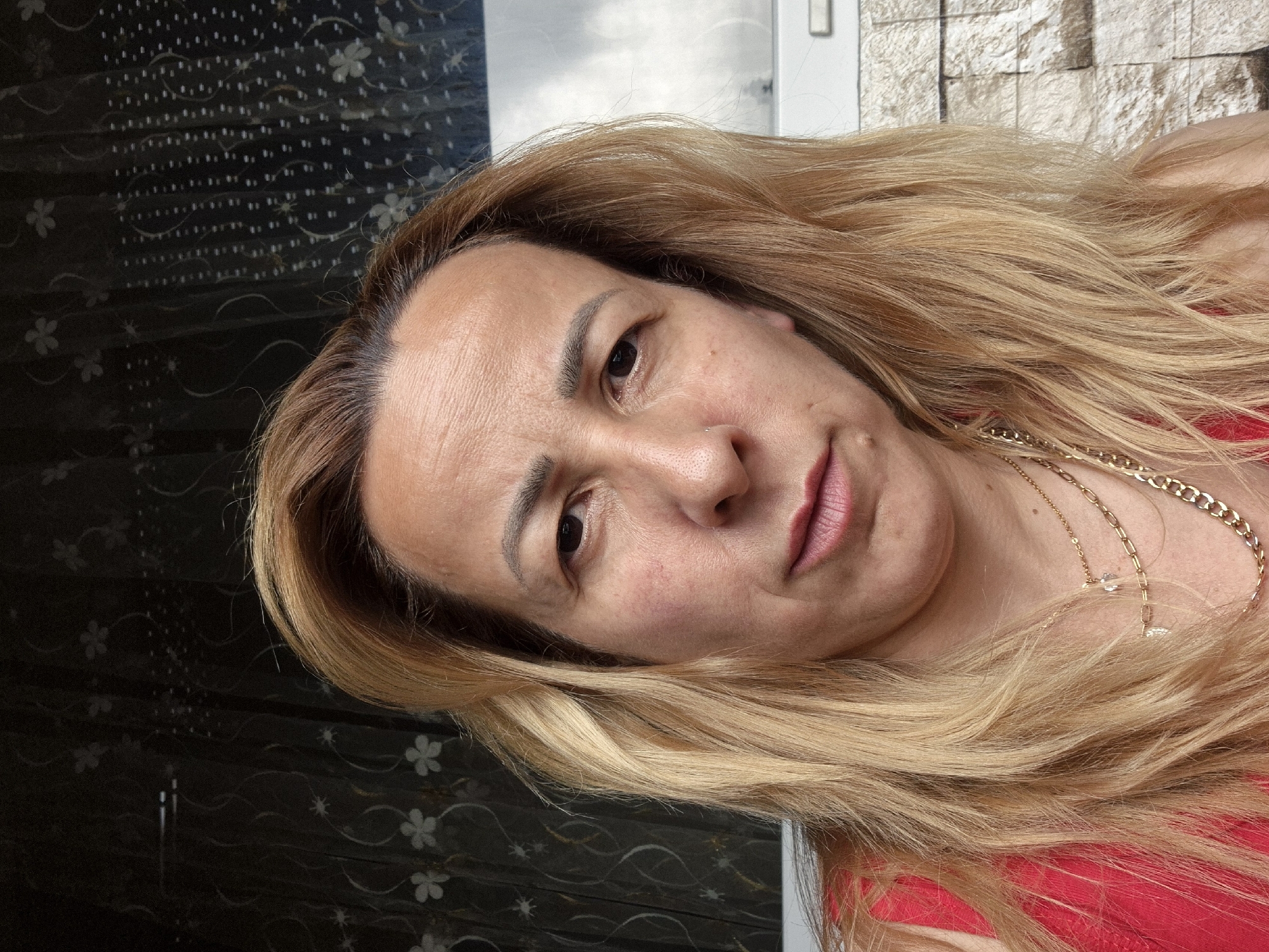 A close-up, angled portrait captures an individual, likely an adult female, with long, wavy blonde hair showing darker roots. The person's face is tilted, looking directly towards the camera with a neutral to slightly serious expression. Dark eyes are visible, and the lips are slightly parted, adorned with a pinkish-red lipstick. A small silver-colored stud is visible as a nose piercing on the right nostril. Fine lines are apparent on the forehead, and there is a subtle reddish discoloration on the left cheek.
The subject is wearing a red garment, possibly a top, and a gold-colored chain necklace with a small, clear or white pendant is draped around the neck. Another, thicker gold-colored chain is also visible.
The background is divided. To the left, there is a dark surface, possibly a curtain or wallpaper, patterned with repeating light-colored flowers and small dots. To the right, a textured wall resembling light-colored bricks or stones is visible. Above this wall, a blurry area of white and light grey suggests either a window or a bright light source. The lighting in the image is bright and even. No specific activity or event is depicted beyond posing for the photograph. There is no visible text.