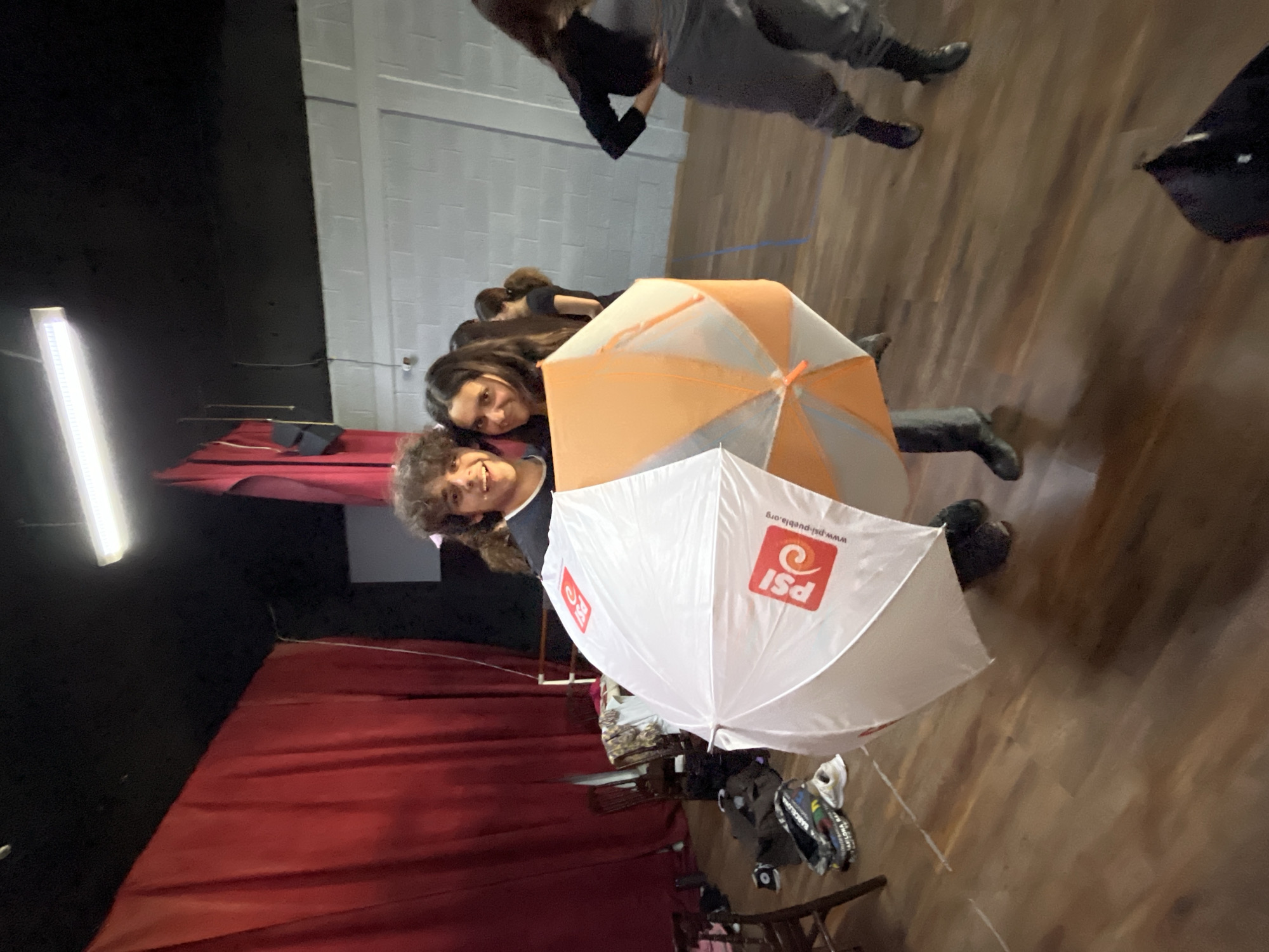 Two young women are smiling at the camera, standing in a room with red curtains, a light fixture, and exposed brick. They are holding two umbrellas: one orange and white, the other white with the PSI logo. The PSI logo reads "www.psi-puebla.org". The location is presumably in Puebla, Mexico, based on the PSI logo. The scene is likely indoors, possibly a theater or studio, given the stage-like setup. Other people are present in the periphery.