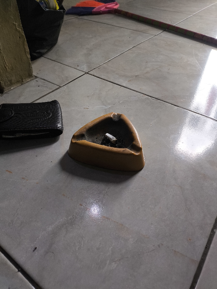 The scene depicts an interior setting on a light grey tiled floor in Cibatu, Indonesia. In the foreground, a triangular yellowish-brown ashtray contains cigarette ash and a single white cigarette butt. To the immediate left of the ashtray lies a closed black wallet with an embossed surface. On the far left, a light-colored vertical structure, possibly a wall base or pillar, is visible, partially obscuring a black plastic bag positioned behind it. In the background, a broom with a reddish-orange and blue head and a striped handle rests on the floor. The tiled floor exhibits visible grout lines and light reflections. No individuals are present in the image.