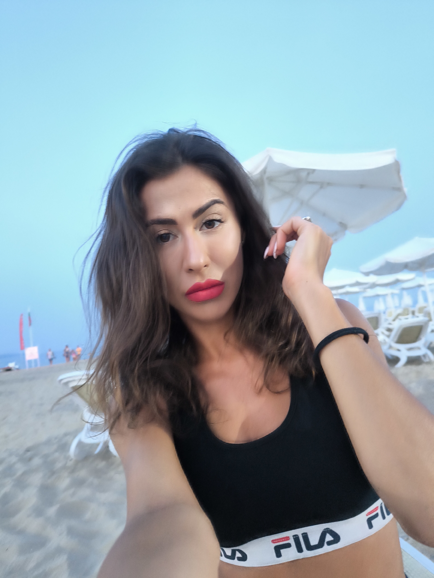 A woman is taking a selfie on a sandy beach. She has dark brown wavy hair and is wearing a black sports bra with a white band that says "FILA". Her lips are painted bright red. In the background, there are white beach umbrellas and lounge chairs, and a few people are walking in the distance. The sky is a clear, light blue. The image appears to have been taken in Nesebar, Bulgaria, possibly near the beach with the iconic red flags and white signs visible.