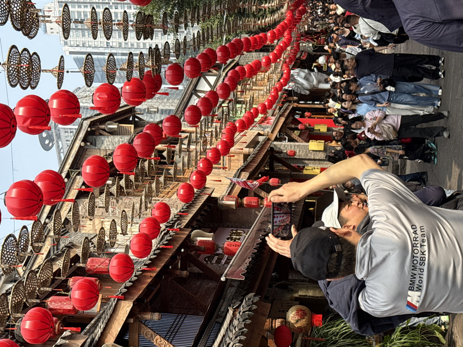 An upward shot shows a street scene adorned with rows of red lanterns. We can see a crowd of people in the background. A person holding a smartphone is taking a picture. Buildings with traditional architecture are visible, contrasting with taller modern structures in the distance. The scene captures a mix of cultural heritage and contemporary life.