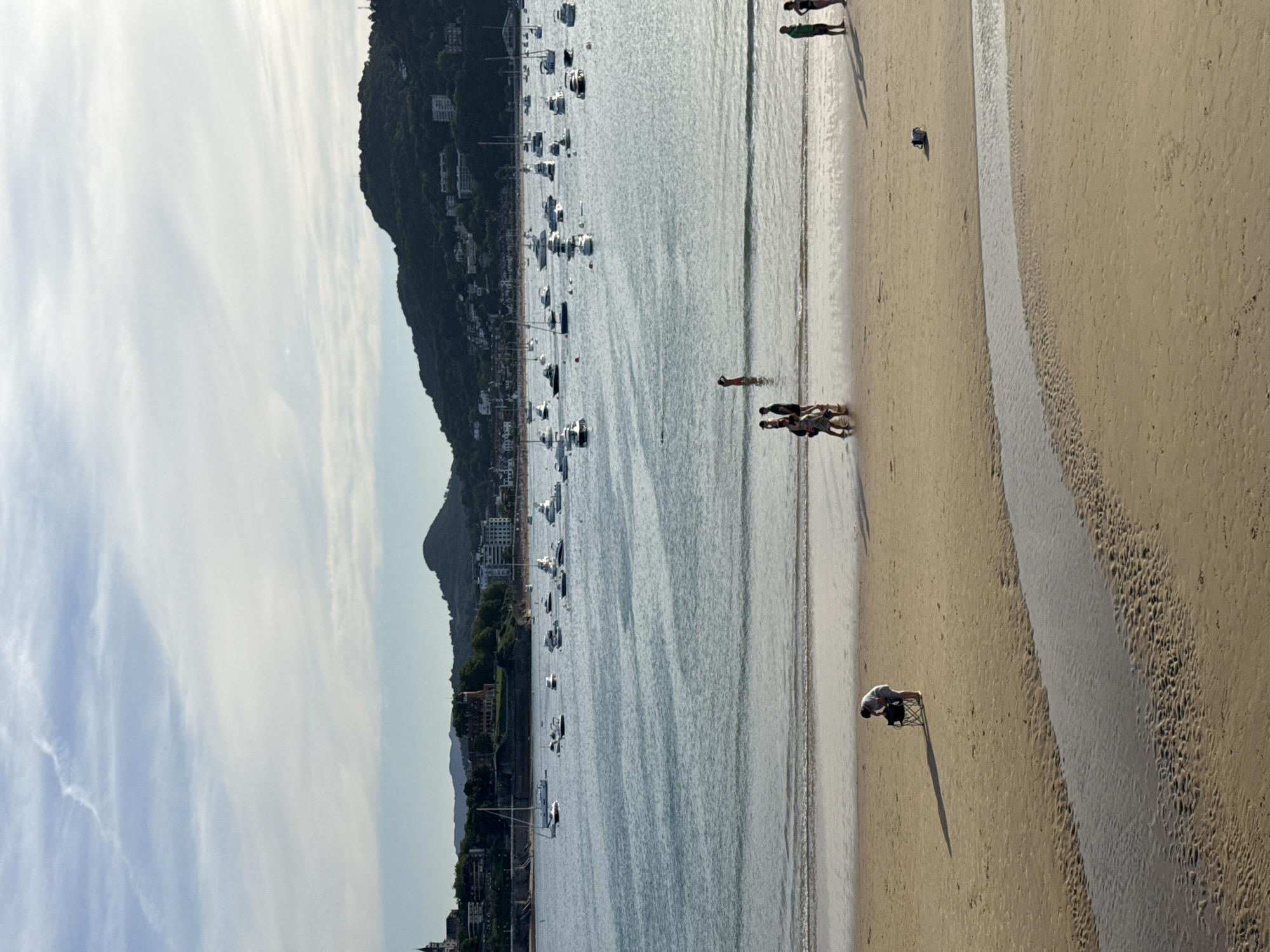 The image depicts a scenic view of La Concha Bay in San Sebastián, Spain, at what appears to be late afternoon. The foreground is a sandy beach with a small stream of water flowing across it. In the mid-ground, several people are visible on the beach. To the left, a person in a wheelchair is looking down at something on the sand. Further to the right, a group of people are walking along the shoreline. In the background, the calm waters of the bay are dotted with numerous boats of various sizes, including sailboats and motorboats. Across the bay, the lush green hills of San Sebastián rise, with buildings and a prominent church spire visible on the slopes. The sky is partly cloudy with soft sunlight casting shadows on the beach.