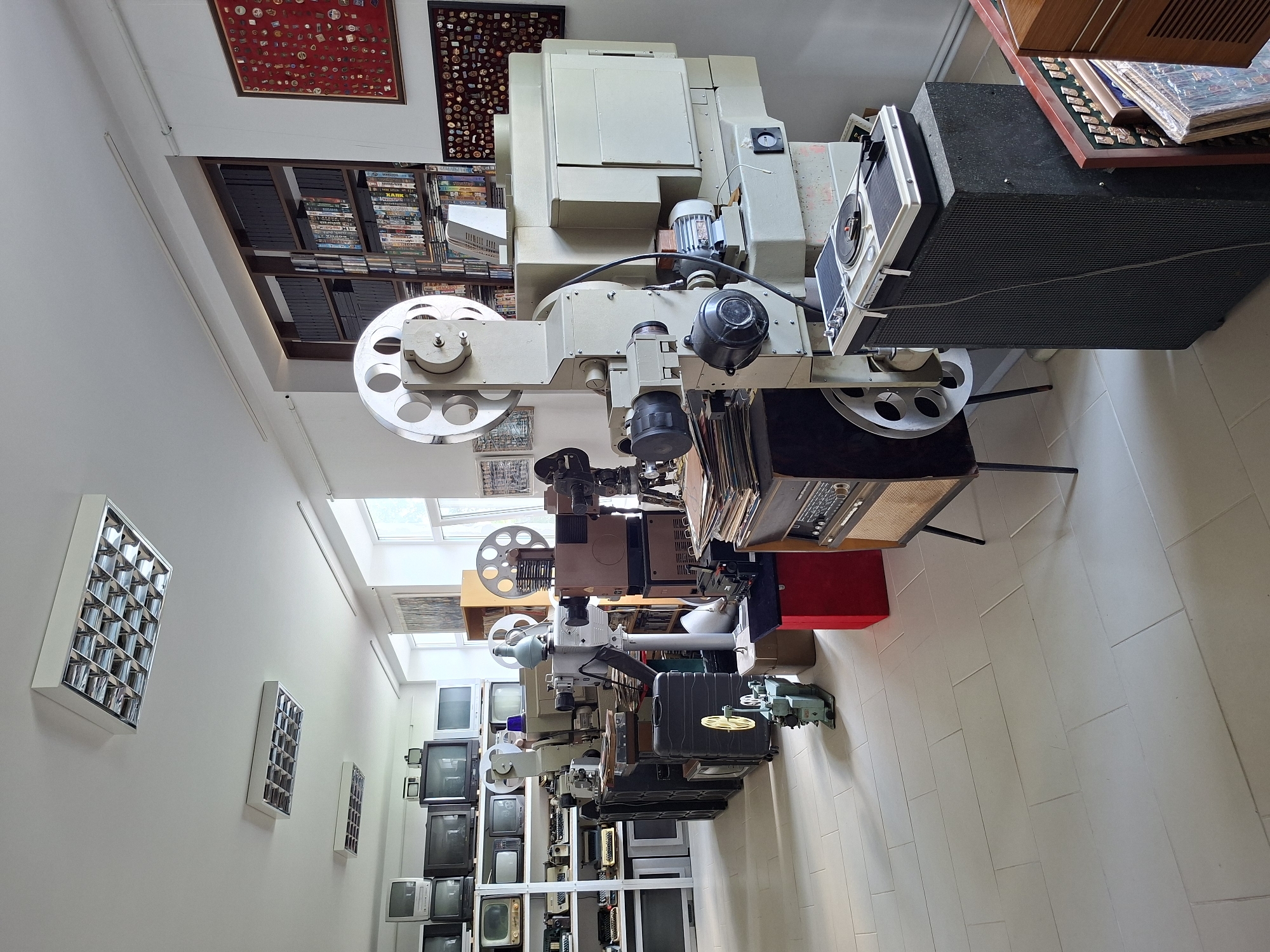 The image depicts a room filled with vintage audiovisual equipment, likely a museum or archive. The room is well-lit with fluorescent ceiling lights. Along the walls are shelves stocked with numerous CRT televisions and video equipment. In the center of the room stand several film projectors of varying sizes, some with reels of film. One projector is particularly large and prominent, with a complex mechanical structure and a large speaker next to it. A turntable sits atop the speaker. To the right, a bookshelf is filled with what appear to be VHS tapes or film canisters. Two framed displays of pins or medals hang on the wall. The floor is tiled with large, pale gray rectangular tiles. The overall impression is one of a preserved collection of historical media technology. This scene is located at the National Museum of Ethnography and Natural History in Chișinău, Moldova.