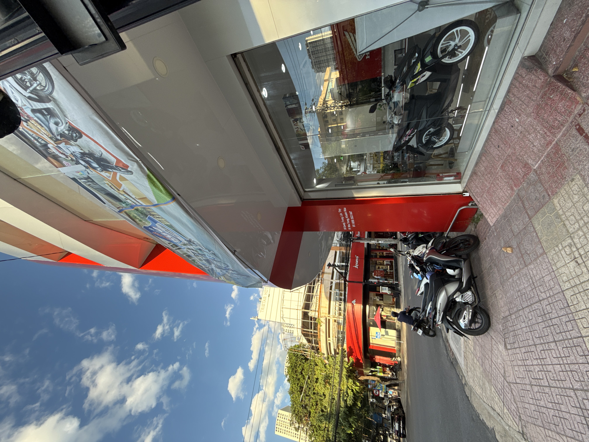 An exterior, ground-level, low-angle shot captures a modern building in Nha Trang, Vietnam, with a bright red facade and large glass windows displaying motorcycles. The sky above is blue with scattered white clouds. In the foreground, a sidewalk made of patterned concrete tiles leads to a ramp. Several motorcycles are parked on the sidewalk and visible through the display windows. The building's architecture features clean lines and a prominent red accent. The street in front of the building is paved and appears to be a public road with vehicles and pedestrians. Some of the text visible on the building and signage indicates it is a Honda dealership. The overall impression is a commercial street scene in an urban environment.