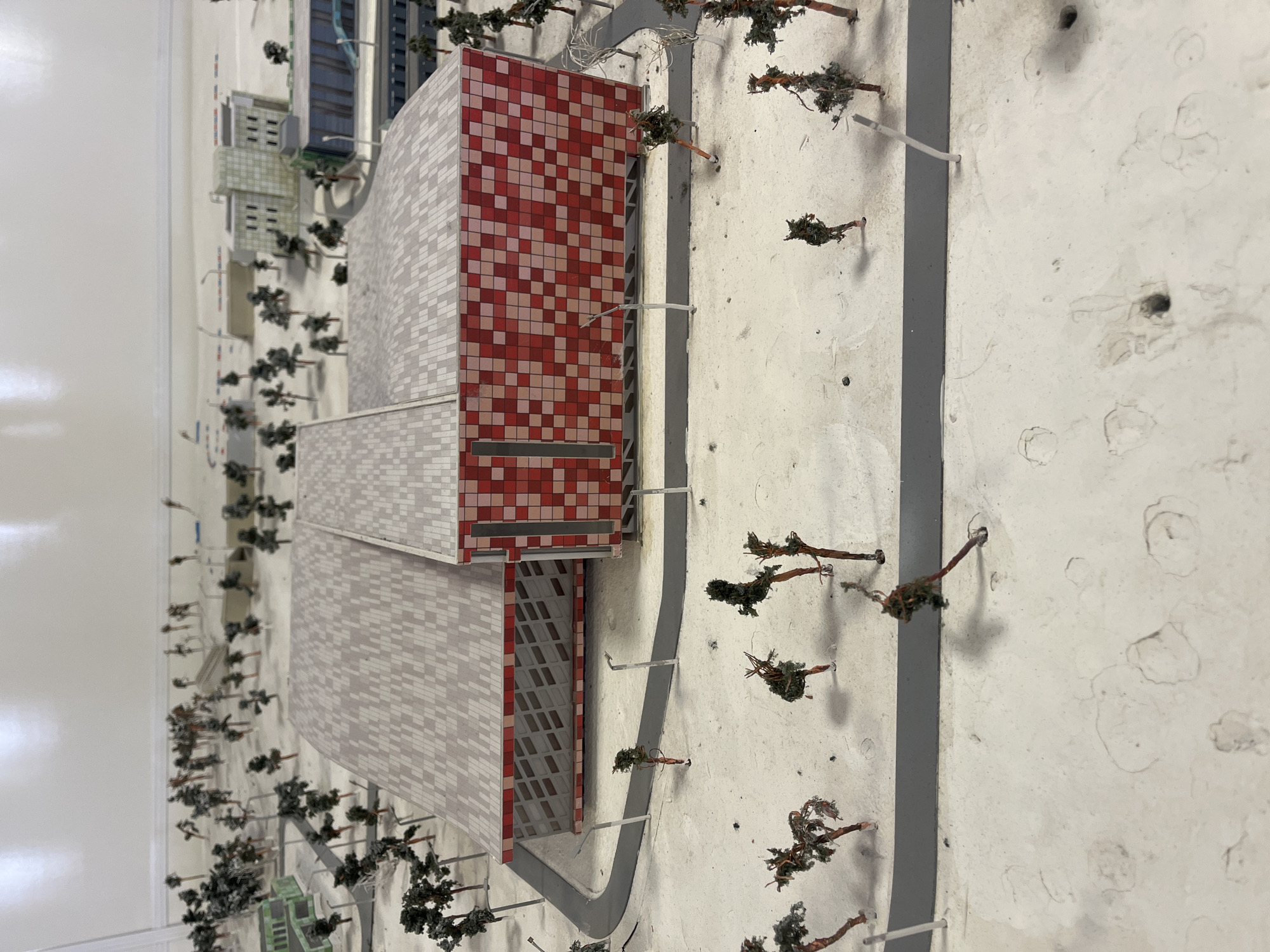 This is a model of a building, potentially located in Krasnoyarsk, Russia. The model features a multi-story building with a facade made of small red and white tiles on part of its surface. The rest of the building is covered with tiles that have the same gray shade. Adjacent to this building is a smaller, generic building. A road, represented by a gray strip, curves around the building. Small, model trees are sparsely scattered along the road and the surrounding white textured surface. Streetlights are also visible along the road. The model is not highly detailed, giving a conceptual overview of the architectural plan. The background is a plain white wall.