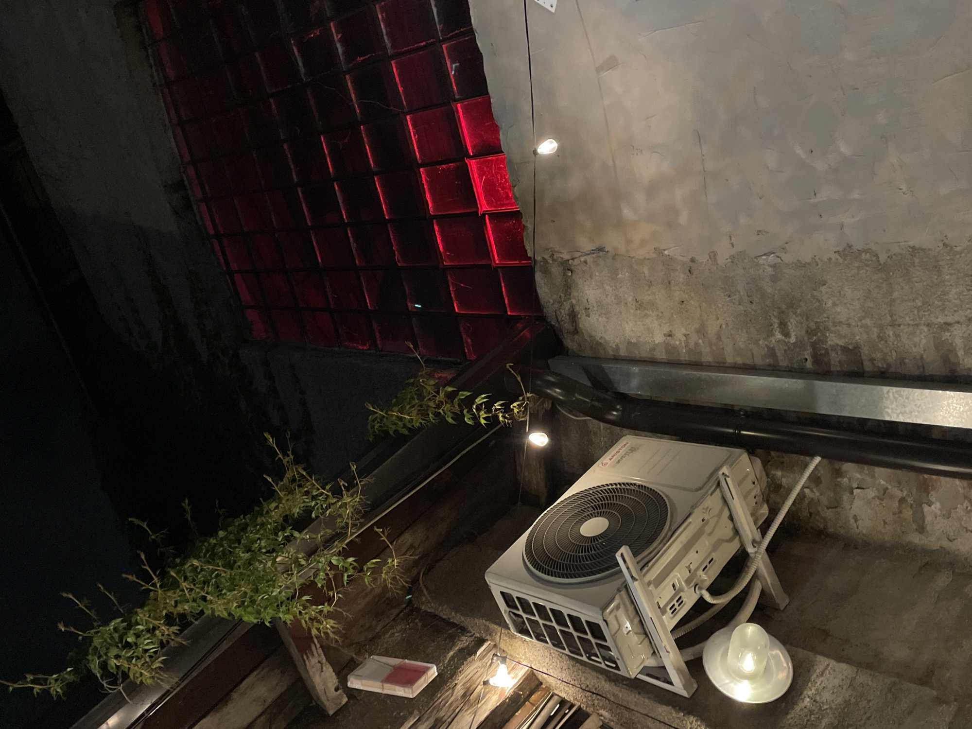 The image depicts a nighttime outdoor or semi-enclosed urban space, viewed from a low, upward-angled perspective. A white Ariston Inverter air conditioning condenser unit is mounted horizontally on a wall bracket in the mid-left. Below it, a white, bell-shaped wall lamp emits a warm, bright light. The lower-left wall exhibits rough textures, including sections of exposed brick and crude plasterwork. Above the AC unit, a dark wooden beam structure, possibly an overhang or ceiling, is visible, from which green, sparsely leaved vines with bare stems trail downwards. A small, rectangular white box with a red segment is affixed to this wooden structure.
In the upper-right quadrant, a prominent grid of square, translucent red glass blocks is intensely illuminated from behind, casting a deep red glow onto the surrounding environment. Below this illuminated section, a vertical black drainage pipe runs down the wall, adjacent to a metallic channel. The wall beneath the red glass blocks features varied plasterwork, appearing rougher and darker on the far left and transitioning to a smoother, lighter-toned surface towards the bottom right. A string of incandescent bulbs hangs diagonally across the upper-middle of the frame, with two bulbs glowing. The upper portion of the image, above the structures and vines, is enveloped in darkness.
The combination of aged architectural elements, modern utility installations, and atmospheric, decorative lighting suggests an outdoor seating area or facade of a commercial establishment, such as a bar or restaurant, in an urban setting. This aesthetic is characteristic of renovated historical districts and popular nightlife areas within the city of Belgrade, Serbia, where old buildings are repurposed for contemporary use. No individuals are present in the scene.