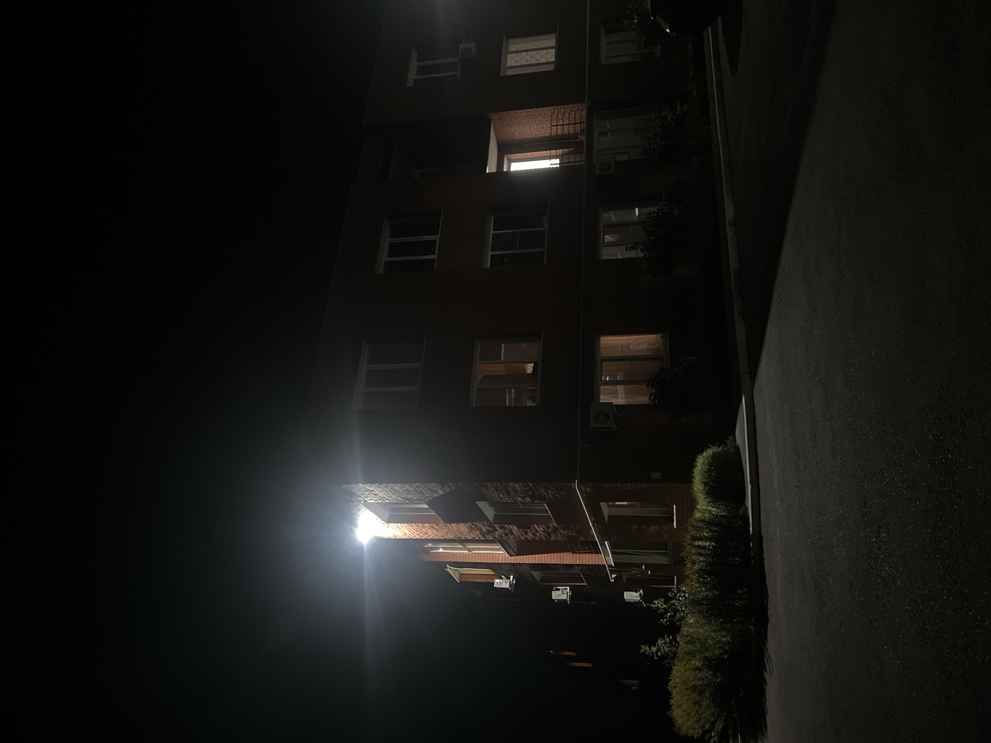 The image captures a brick apartment building at night in the city of Nezhinka, Russia. A bright overhead light illuminates the corner of the building, casting a strong glare. The building has multiple windows, some of which are lit from within, indicating occupancy. Several outdoor air conditioning units are visible mounted on the exterior walls. In front of the building, a well-maintained hedge runs along a paved area. The foreground shows a dark asphalt surface, likely a road or parking lot, with shadows cast by the building and the hedge. The overall scene is dark and atmospheric, typical of a residential area at night.