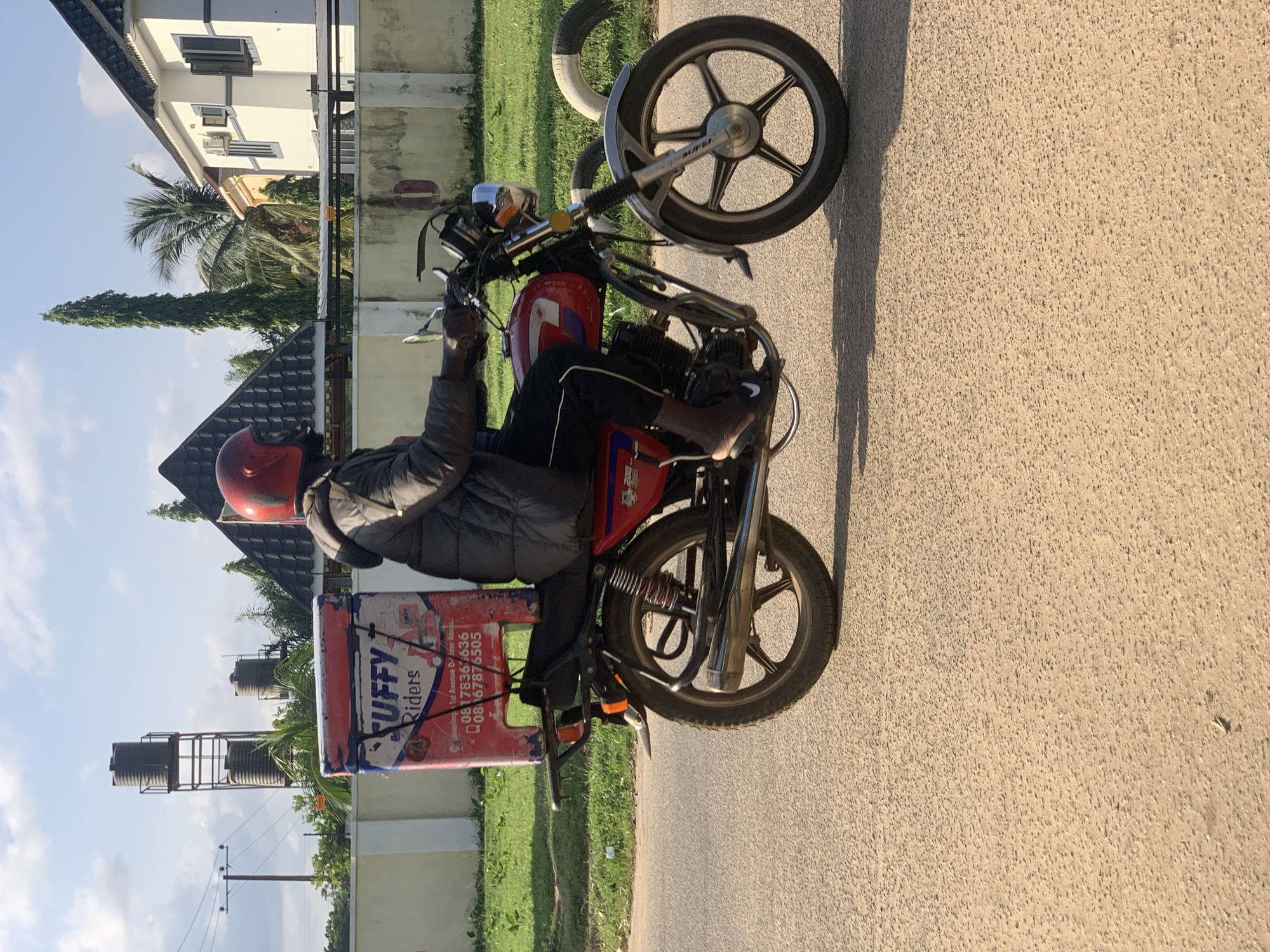 A person wearing a grey puffer jacket and a red helmet rides a red motorcycle on a paved road in Abuja, Nigeria. The motorcycle has a large, red, box-shaped container attached to the back, with the word "STUFFY" and "Riders" written on it. The container also displays phone numbers. In the background, there are buildings, including a white house with dark trim, a black-tiled roof, and multiple windows. To the left of the house, there is a tall, slender tree and palm trees. Further in the background, there are water tanks on a metal structure and power lines. The ground is asphalt, and there is a strip of green grass on the side of the road. The sky is blue with scattered white clouds.