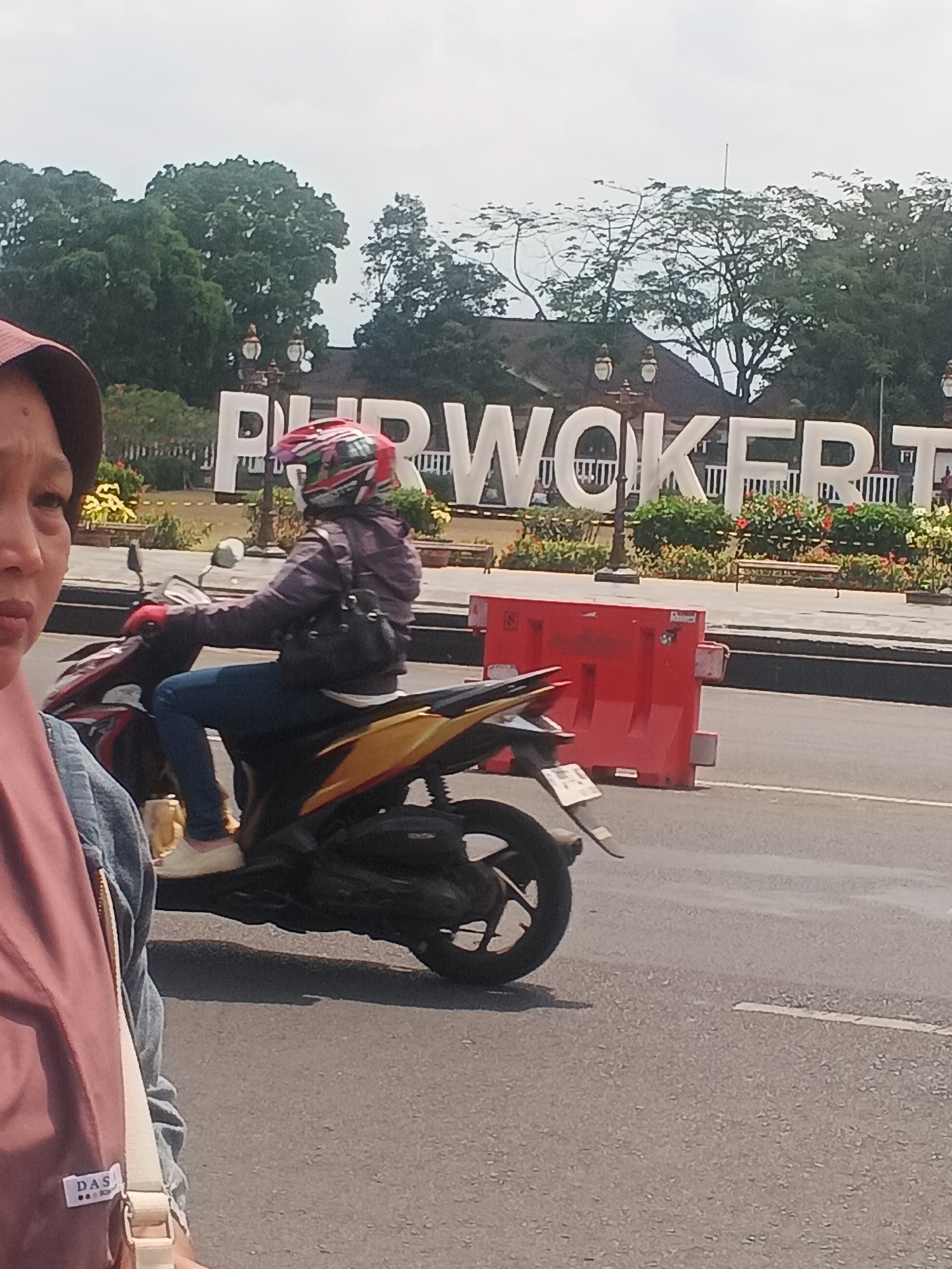 A person wearing a helmet and a purple jacket rides a red and yellow motorcycle. The person is on a street with a large white sign that says "PURWOKERTO" in the background. There are trees and bushes behind the sign. A red barrier is on the right side of the road. In the foreground, a person wearing a pink headscarf and a blue denim jacket is partially visible. The person is holding a bag with a "DAS" label. The image appears to be taken in Purwokerto, Indonesia, likely at a tourist attraction or landmark featuring the city's name.