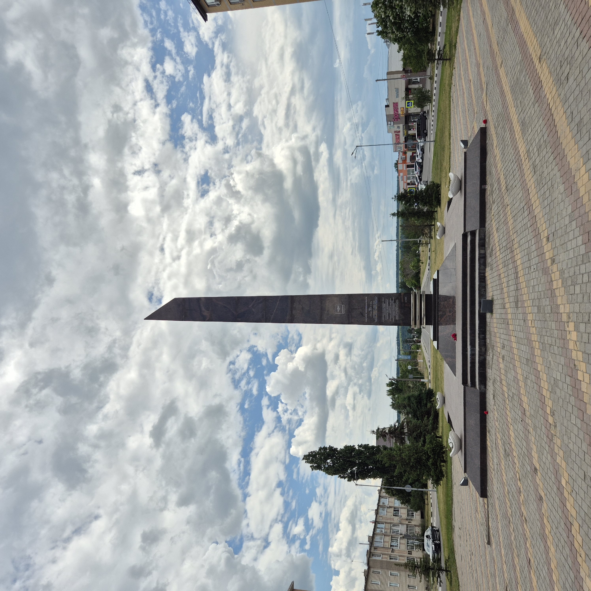 This is a frontal, eye-level, wide-angle photograph of the obelisk monument located in the city of Liski, Russia. The monument is a tall, slender, dark obelisk with a sharply angled top, positioned centrally in the frame. It stands on a multi-tiered, dark stone base with steps leading up to it. Two white, boat-shaped planters are visible on either side of the base, and a single red rose lies on the lower steps of the monument.
The monument is situated in a paved plaza with a distinctive pattern of reddish-brown and yellow rectangular paving stones. In the background, to the left, is a multi-story apartment building constructed of light-colored brick. To the right of the monument, a street is visible, lined with trees and shops. One shop has a sign that reads "КОСМЕТИК". Cars are parked along the street and a dark sedan is driving on the road. The sky is filled with dramatic, fluffy white and grey clouds against a backdrop of blue sky. Several tall, dark green evergreen trees are planted around the plaza and in the distance. The overall scene depicts a public space, likely a memorial or park, in a modern urban or suburban setting.