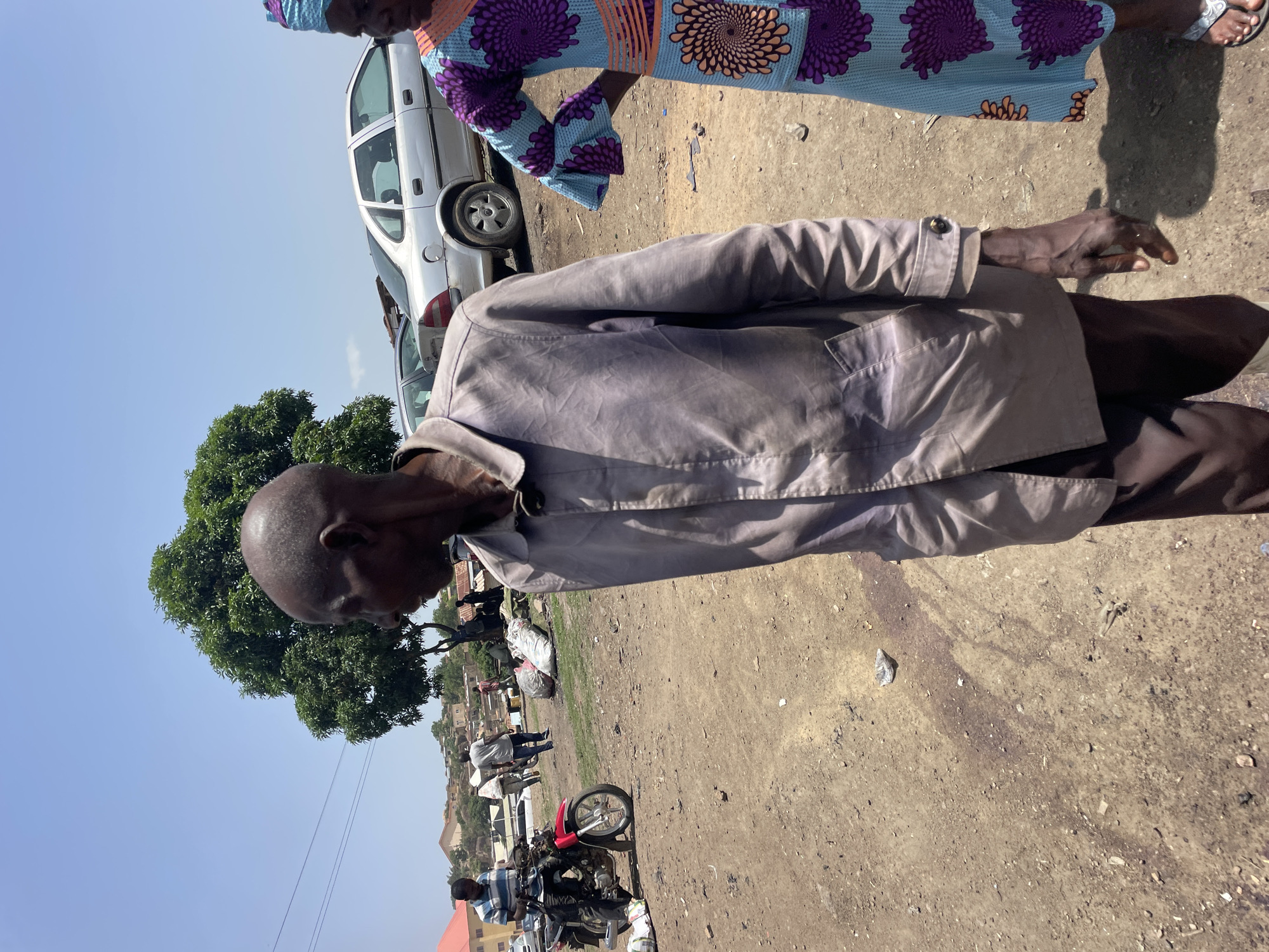 The image captures a scene in Jos, Nigeria, featuring an older man wearing a light-colored jacket. He stands on a dusty, dirt surface scattered with debris. Behind him, a silver car is parked, and a person wearing a bright patterned dress is partially visible. In the background, a tree provides foliage, and further away, a red motorcycle and several people are visible, suggesting a marketplace or gathering area. The sky is clear and blue. The image is taken at ground level, with a slight tilt.