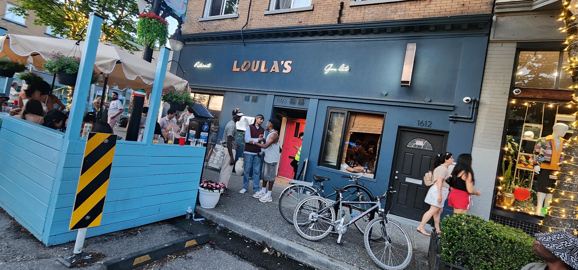 The image shows an outdoor seating area in front of a building with "LOULA'S" signage. Several people are seated at tables in the blue outdoor structure, some of whom appear to be dining. In front of the building, three men are standing and interacting, two of them facing each other. Two bicycles are parked on the sidewalk. A storefront with a mannequin and decorative lights is visible to the right of the building. The building itself has windows, doors, and security cameras. The scene appears to be in a city environment with trees and streetlights visible.
