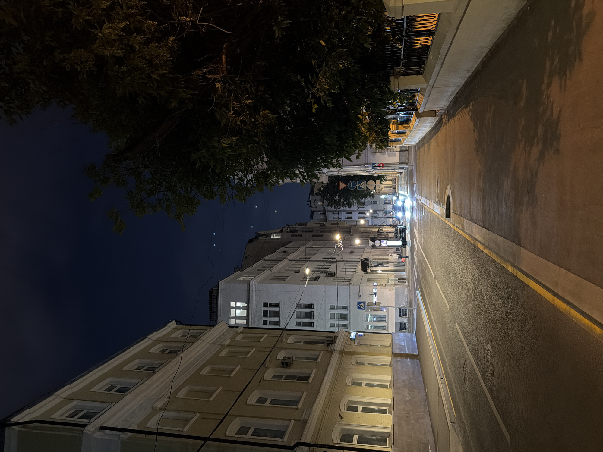 The image depicts a nighttime street scene in Moscow, Russia. On the left side of the street, a light yellow building with multiple windows lines the sidewalk. Across the street, a series of multi-story buildings with white facades and numerous windows are visible. The street itself is paved and illuminated by streetlights, casting shadows from trees and fences onto the sidewalk on the right. A few cars are on the road, their headlights on. A single person stands on the sidewalk near a parked car. The sky above is dark blue, with some faint stars visible. The environment appears to be an urban residential area.
