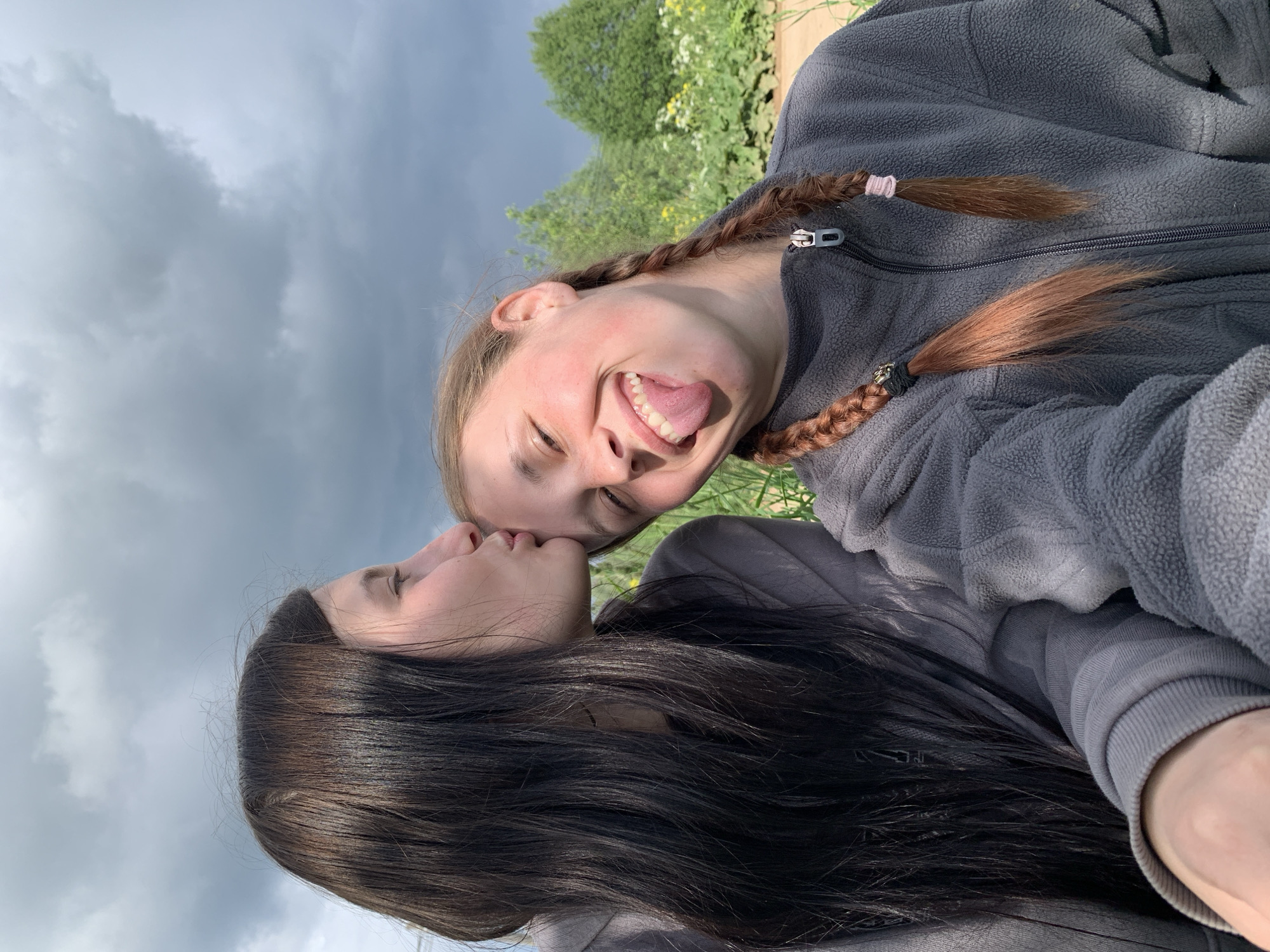 Two women are closely positioned, possibly kissing. One woman has dark hair, and the other has light hair styled in braids. The light-haired woman is sticking out her tongue. They both wear gray fleece jackets. The background includes a cloudy sky and some greenery, possibly vegetation in a field. The image appears to be taken outdoors, possibly on a cloudy day. There is no indication of the location being Фоки, Russia, in the image.