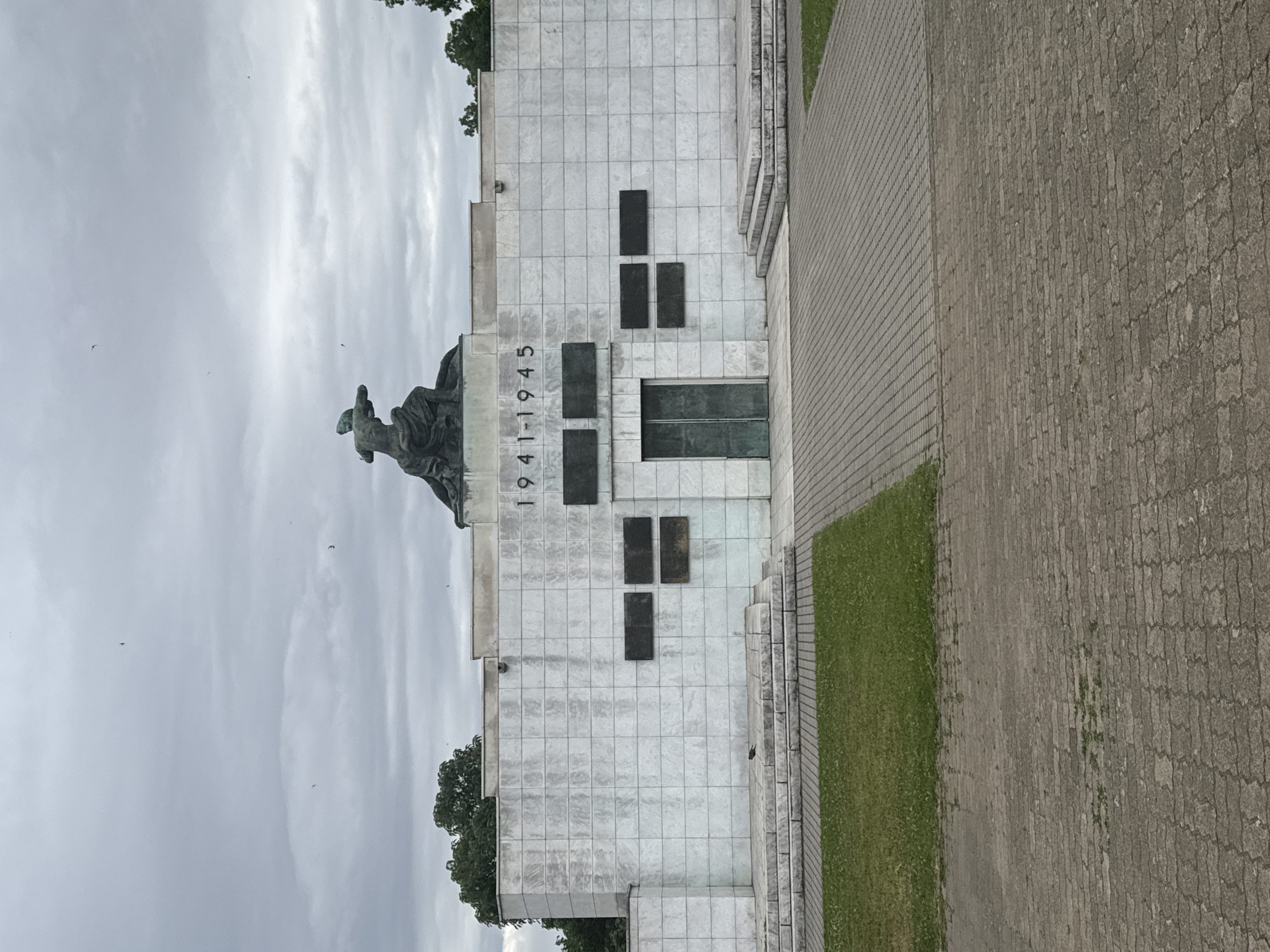 The image shows the Memorial Ossuary of the Fallen for the Fatherland in the city of Subotica, Serbia. The ossuary is a large, white marble structure with a bronze sculpture of a kneeling figure on top. The dates "1941-1945" are inscribed on the facade. There are several dark rectangular plaques on the wall, and a set of double doors in the center. To the left and right of the doors are tiered stone steps. In front of the structure is a paved walkway and a grassy area. The sky is overcast, and there are a few birds in the sky. A lone bird is perched on the steps to the left.