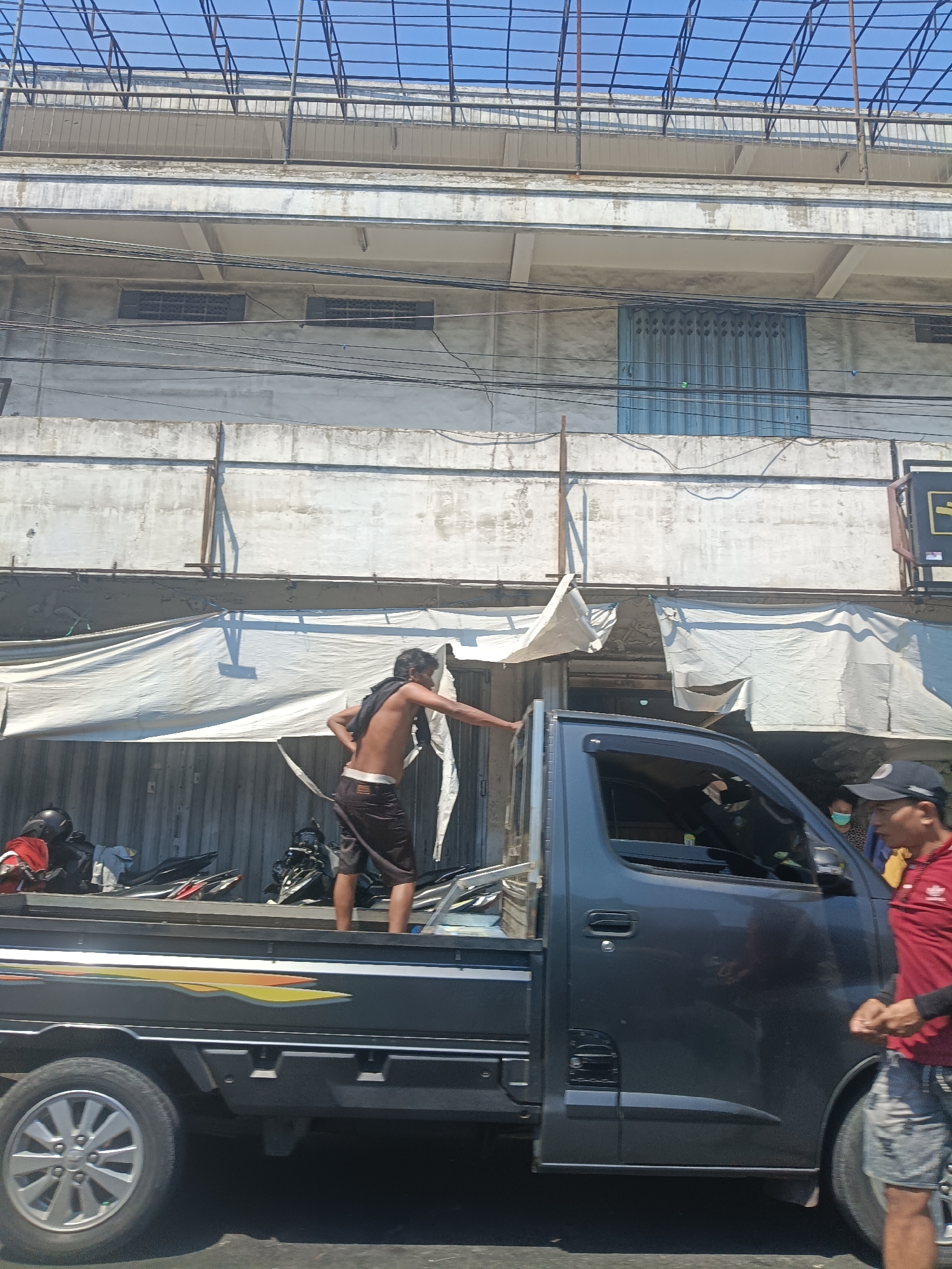 In Surabaya, Indonesia, on a sunny day, a shirtless man wearing dark shorts stands in the bed of a dark grey pickup truck. He has a black cloth draped around his neck and is reaching out with his right hand towards the passenger side of the truck. Several motorcycles are visible in the truck bed. In the background, a building with a white facade and rows of windows is visible, with a corrugated metal facade underneath a white tarp. A person wearing a red shirt and a baseball cap is walking past the truck on the right side of the image, while another person wearing a face mask is visible inside the truck. The sky is clear and blue.