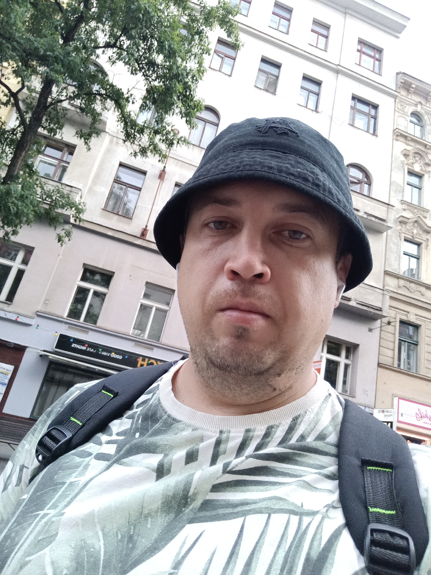 In Prague, Czechia, a man is photographed from the chest up. He is wearing a black bucket hat, a white t-shirt with a green palm leaf pattern, and a black backpack. He has a faint stubble and is wearing white wireless earbuds. The background consists of an old building with multiple windows and a tree with green leaves on the left. A sign with text is visible on the building behind him.