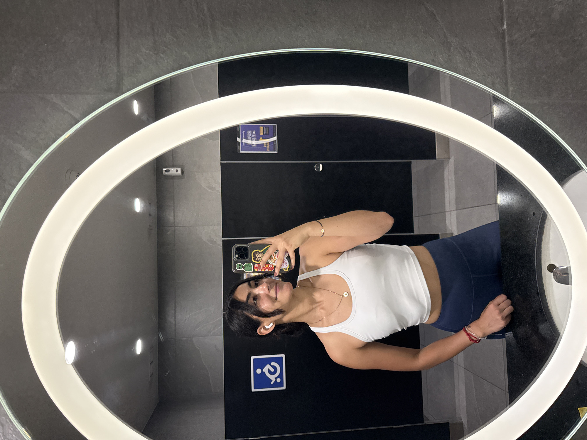 A young woman stands in a bathroom, likely a public restroom. The scene is dominated by a large, round, lighted mirror reflecting the woman and the bathroom interior. The woman is taking a selfie with her phone. She is wearing a white tank top and blue leggings. The background reveals gray tiled walls, black stall doors, and a sink. A handicap symbol is visible on one of the stall doors. A small sign is also visible on one of the stall doors. The location of the bathroom is in Puebla, Mexico.