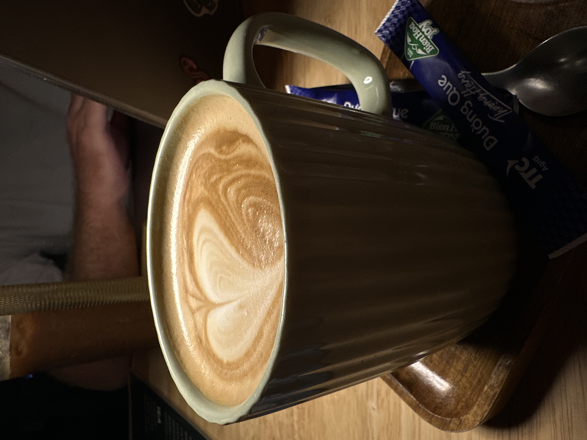 A close-up shot depicts a large, light green ceramic mug filled with latte art, placed on a wooden surface. Adjacent to the mug is a silver spoon and two blue and white sugar packets with Vietnamese writing. A person's arm is visible on the left side of the frame. The scene is likely indoors, possibly in a café or restaurant, based on the presence of the latte and sugar. The sugar packet identifies a brand name in Vietnamese that is consistent with products available in Vietnam. Given the Vietnamese writing on the sugar packets, it's plausible the location is in Bac Nha Trang Ward, Vietnam.