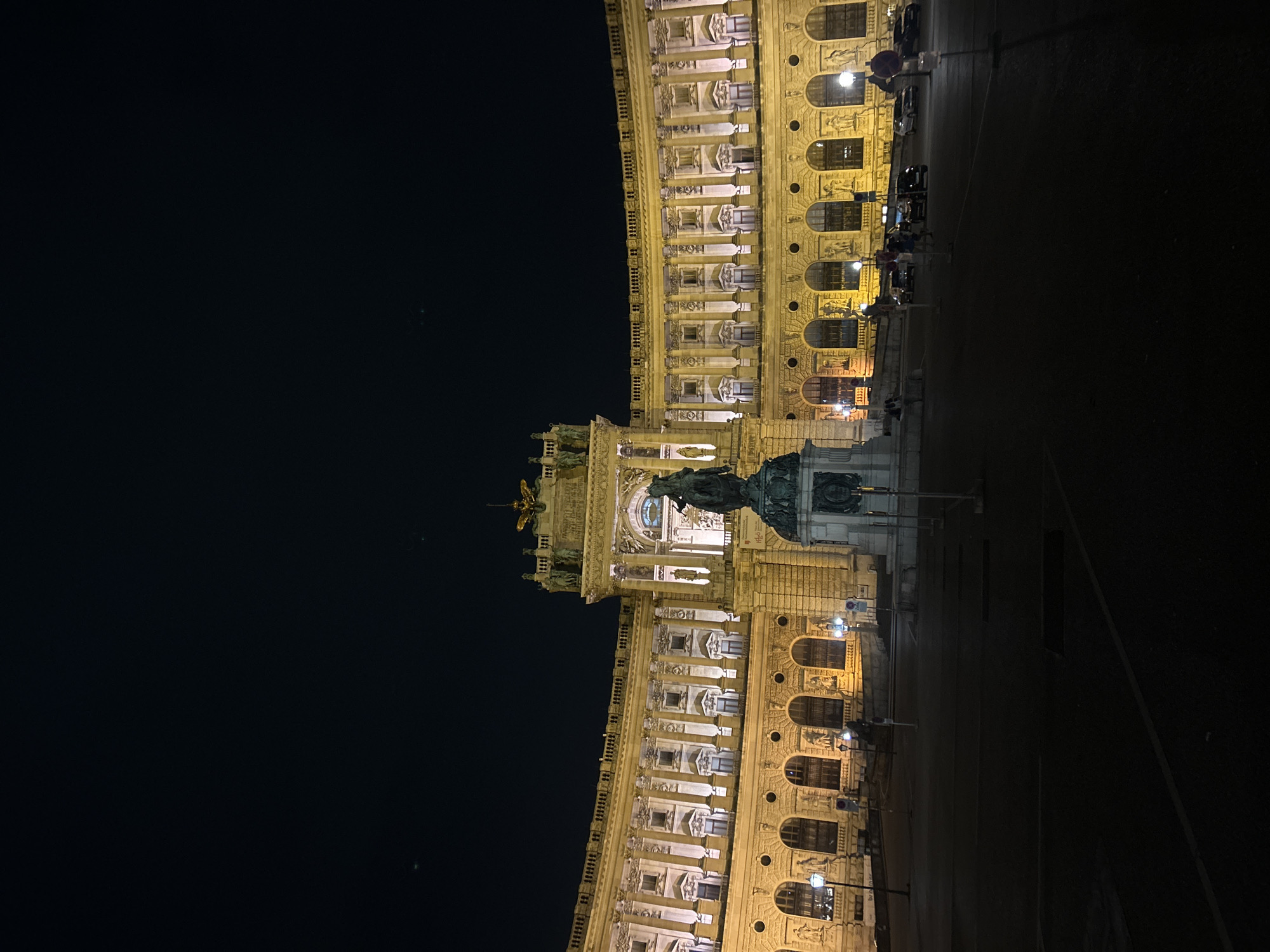 The image shows a nighttime view of the Hofburg Palace in Vienna, Austria. The grand, ornate facade of the palace is illuminated with warm, yellow lights, highlighting its architectural details, including rows of arched windows and classical columns. In the foreground, a large equestrian statue stands prominently in a courtyard. The sky above is dark, indicating it is late at night or early in the morning. Several street lamps are visible along the facade, casting pools of light onto the cobblestone ground. A few cars are parked in the distance, and a couple of figures are seen walking or standing in the courtyard. The overall scene conveys a sense of grandeur and historical significance.