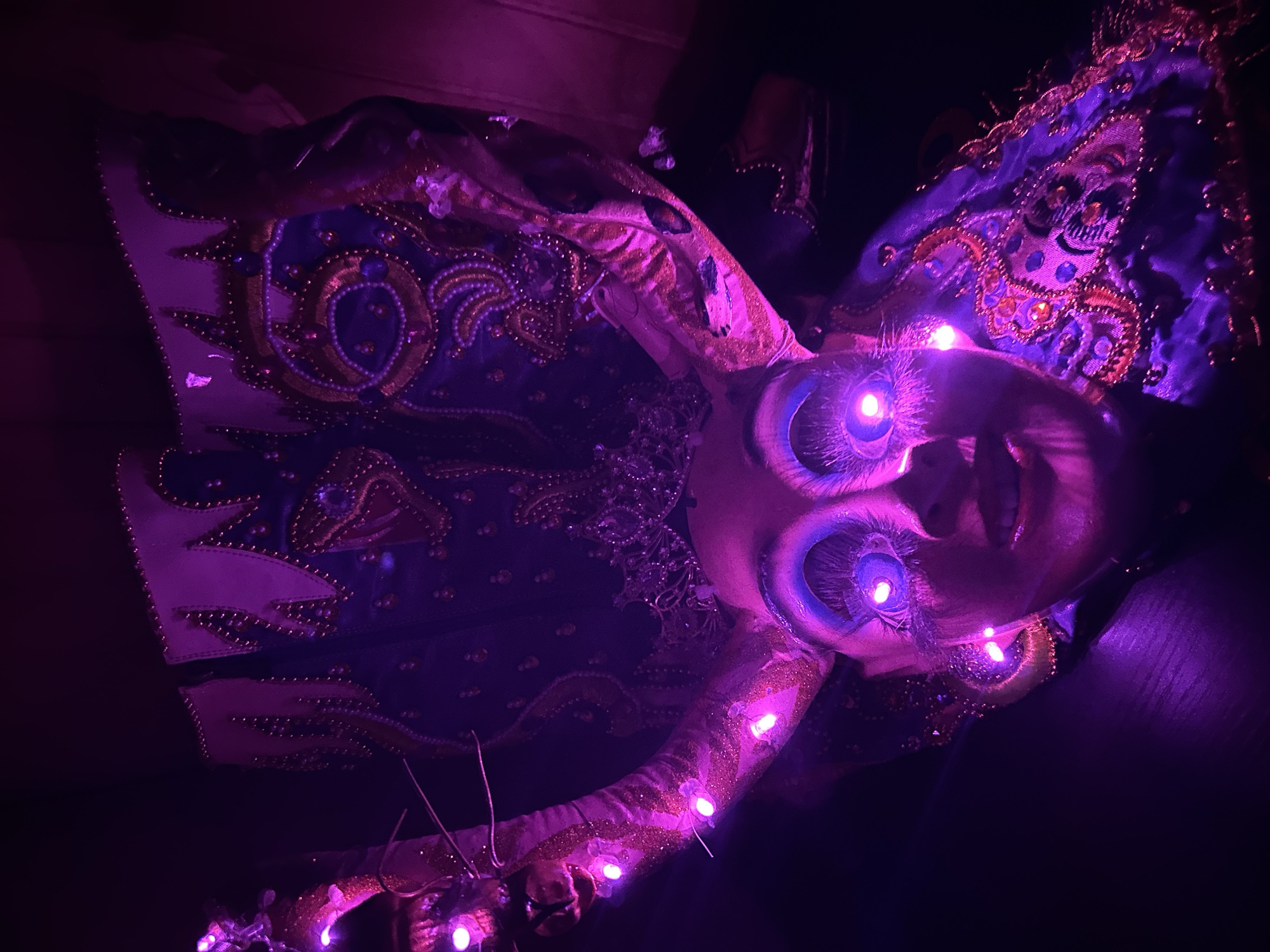 A person in a costume illuminated by purple lighting, possibly in Arica, Chile. The costume features ornate embroidery and detailing, with light-up eyes and lights strung across the costume. The setting appears to be low light.
