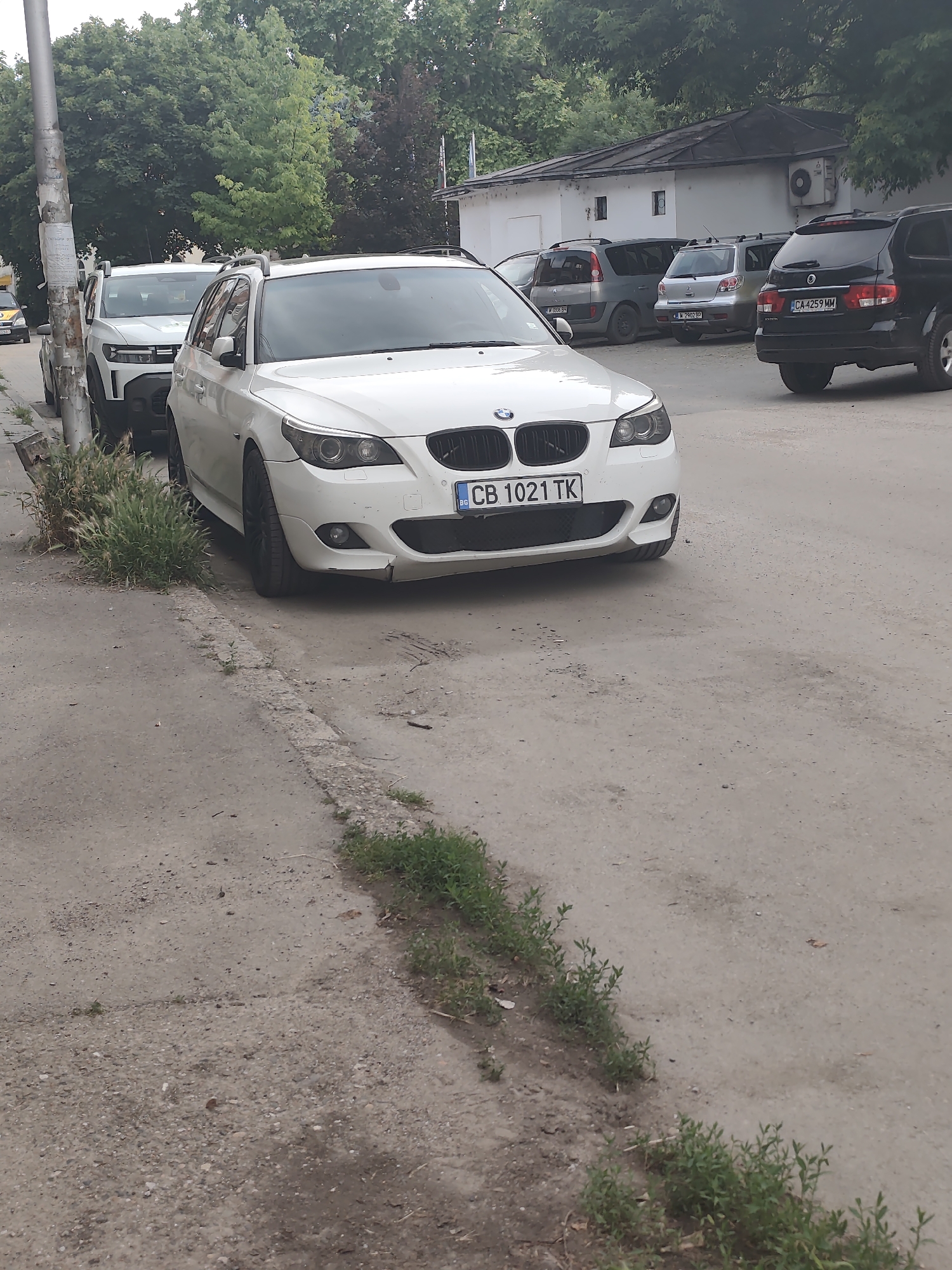 The image displays an outdoor scene in Lom, Bulgaria, featuring several parked vehicles and a building under a tree canopy during daylight. In the foreground, a white BMW 5 Series Touring (E61) is parked on an unpaved shoulder of dirt and gravel, facing the viewer. Its front license plate displays 'CB 1021 TK', and minor damage is visible on the lower right side of its front bumper. Immediately behind the BMW, a white Dacia Duster is partially visible, parked on the same shoulder next to a metal utility pole covered with various faded papers.
Beyond these vehicles, in a separate unpaved parking area, several other cars are parked. These include, from left to right: a grey Renault Scenic (first generation) with the license plate 'M 0089 BH', a silver Mitsubishi Outlander (first generation) displaying license plate 'M 2902 BP', and a black SsangYong Rexton with the license plate 'CA 4259 MM'. Behind these parked vehicles stands a white, single-story building featuring a dark-colored roof, two small rectangular windows, and an exterior air conditioning unit mounted on its wall.
The ground surface in the immediate foreground consists of uneven dirt, gravel, and fragments of concrete, with small patches of green vegetation and weeds growing along a broken concrete curb. Large green trees form a dense background, obscuring the horizon. No people are present in the scene.