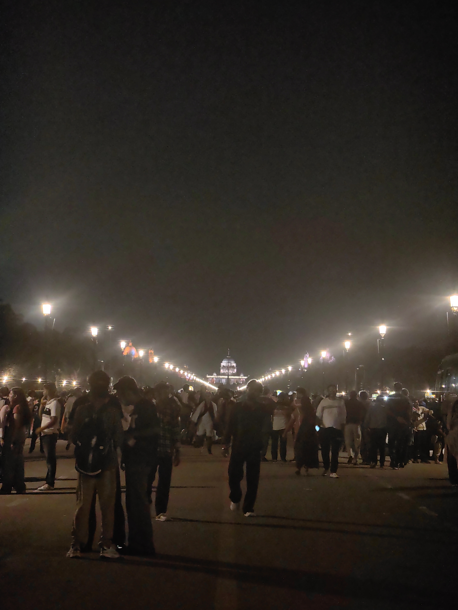 The image depicts a nighttime scene on a wide road in New Delhi, India, likely the Kartavya Path (formerly Rajpath), with a large crowd of people present. The road is illuminated by rows of streetlights, casting a warm glow and creating a sense of depth. In the distance, at the end of the illuminated avenue, a large building with domes and pillars is visible, which appears to be Rashtrapati Bhavan. Numerous individuals are walking in various directions along the road, some in groups and others individually. They are dressed in casual attire, and their faces are largely indistinct due to the low light conditions and the distance. The overall atmosphere suggests a public gathering or a leisurely evening stroll by many people in a prominent location in the capital city.