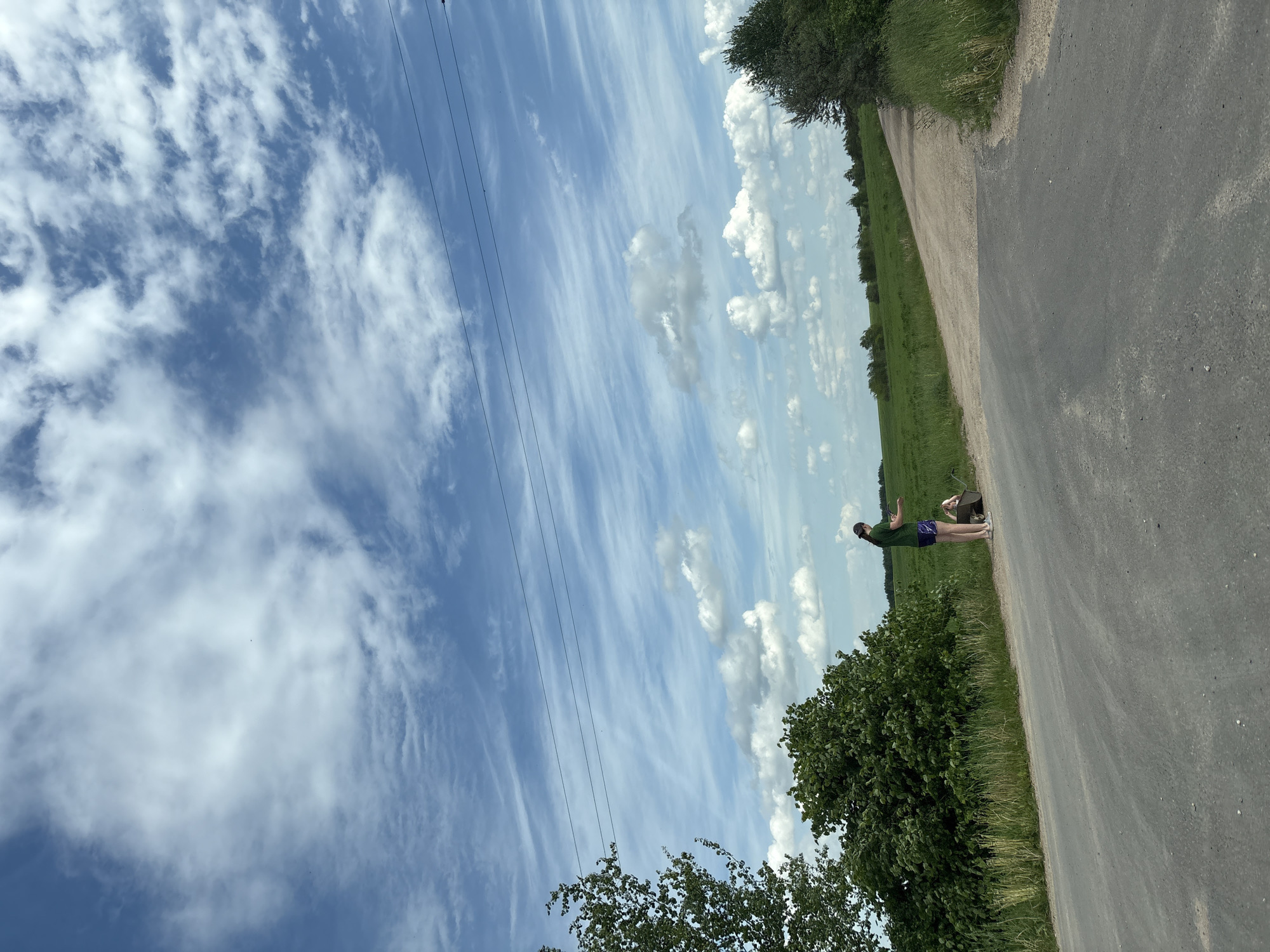 A horizontal photograph shows a paved road in Пярэжырскі сельскі Савет, Belarus. A person stands on the road with a wheelbarrow. Green fields and trees line the road. The sky is blue with streaks of cirrus clouds. Power lines are visible in the sky.