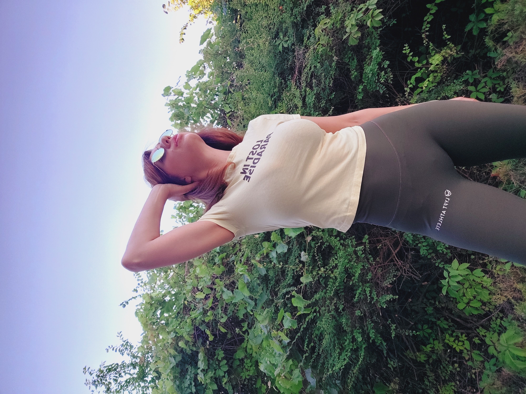 A low-angle, medium shot shows a woman posing outdoors in front of a dense green bush. The sky above is a gradient of pale purple to light blue. The woman, with auburn hair and wearing reflective sunglasses, has her left arm raised, with her hand resting on the back of her head. She is looking upwards and to her right. She is wearing a light yellow t-shirt with black text that reads "LOST IN PARADISE" in reverse and black leggings with white text on the left thigh that reads "FLY ATHLETE" in reverse with a small logo. The foliage is lush and appears to be in a park or natural setting. The time of day appears to be late afternoon or early evening, given the lighting and sky color. The location is the city of Sozopol, Bulgaria.