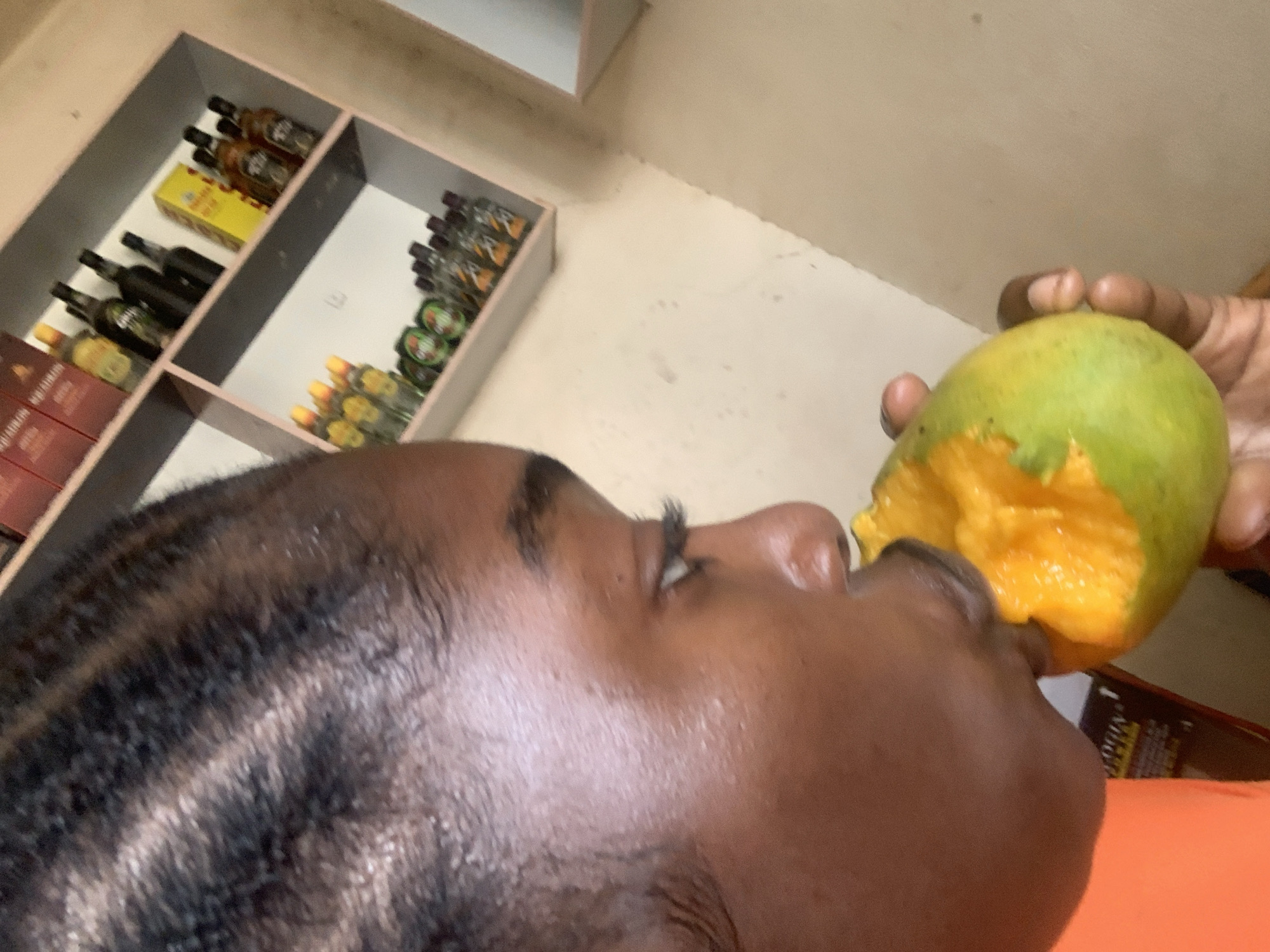 A person is eating a mango. The individual has dark skin and short, dark hair. The mango is green and yellow with orange flesh where it has been bitten. A hand holds the mango. In the background, a shelf holds multiple small bottles of liquid, possibly alcohol. The location is likely a shop in Kabong, Nigeria.