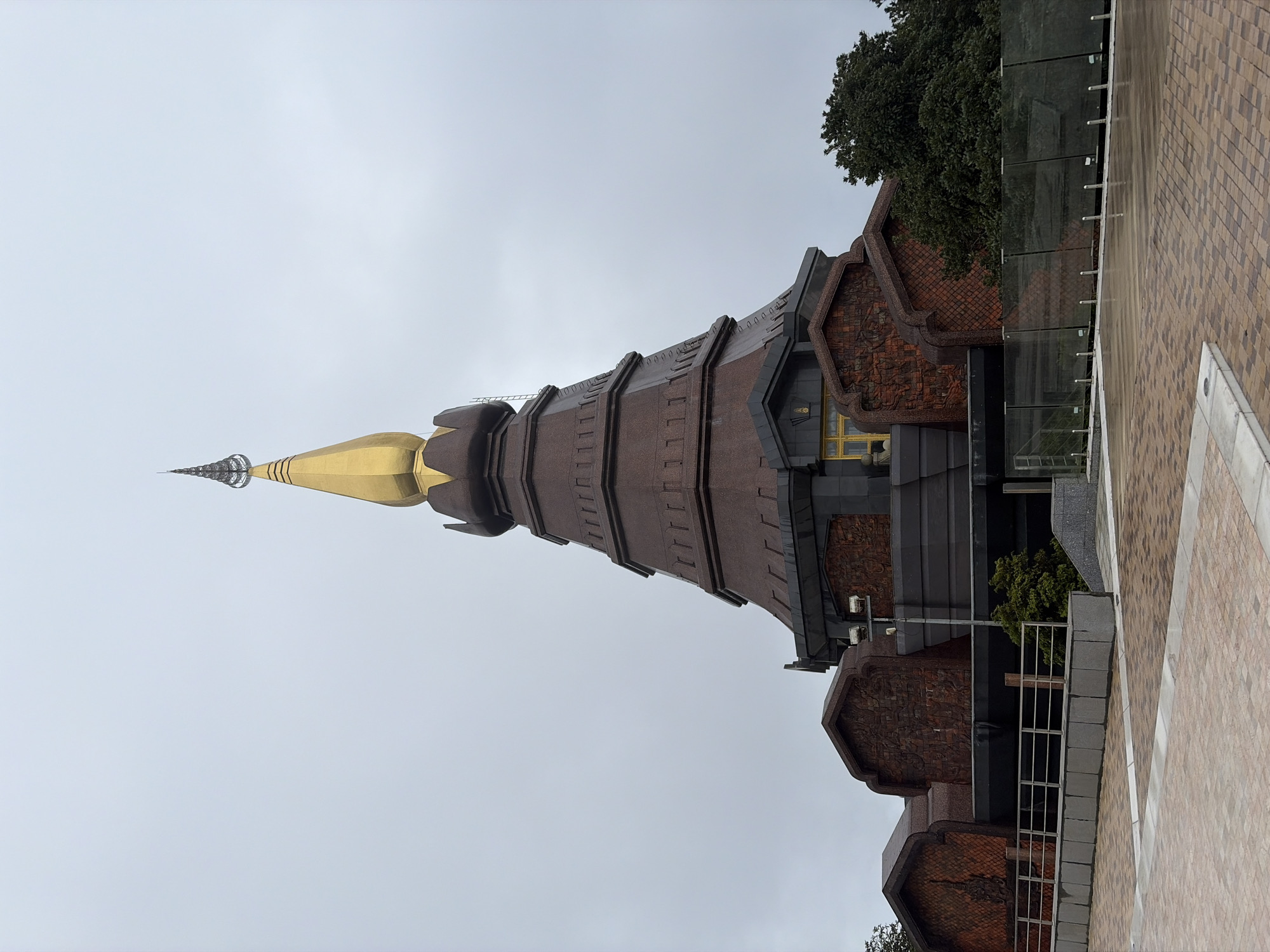 A wide-angle, eye-level shot captures the Phra Mahathat Naphamethanidon, a prominent Buddhist chedi located on Doi Inthanon in Chom Thong District, Thailand. The structure, predominantly made of dark brown stone and accented with a golden spire, dominates the frame under a muted, overcast sky. The chedi is tiered, with a distinctive conical shape. The lower sections of the chedi are adorned with intricate, reddish-brown patterned panels. A metal railing and a low stone wall are visible in the foreground, separating a paved area with a brick pattern from the grounds surrounding the chedi. Green trees are visible to the right of the structure, and part of a dark-colored fence with glass panels lines the middle ground. The ground in the foreground appears wet, suggesting recent rain. No people are present in the image.