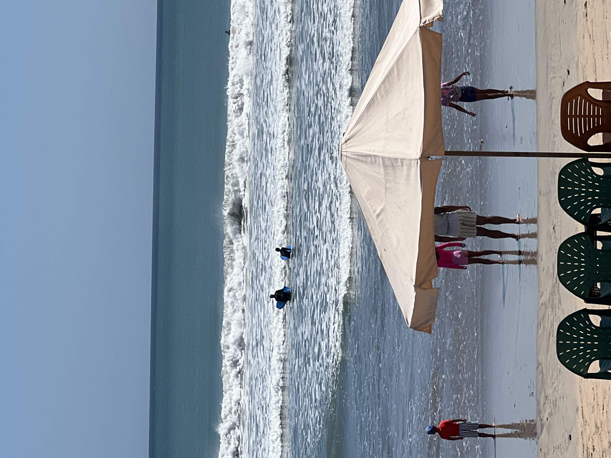 The image depicts a beach scene in Kuta, Indonesia, with several people engaged in various activities. In the foreground, two dark green beach chairs are visible on the sand, with a partially obscured light-colored umbrella providing shade. To the right of the umbrella, a person with dark hair and a colorful top is walking towards the water. Further to the left, a child wearing a red shirt and striped shorts stands facing the sea. In the middle ground, a group of people are in the water. Two individuals are seen with bodyboards, preparing to surf, while another person appears to be floating on a bodyboard. The ocean exhibits moderate waves and white foam. The sky is clear and blue.