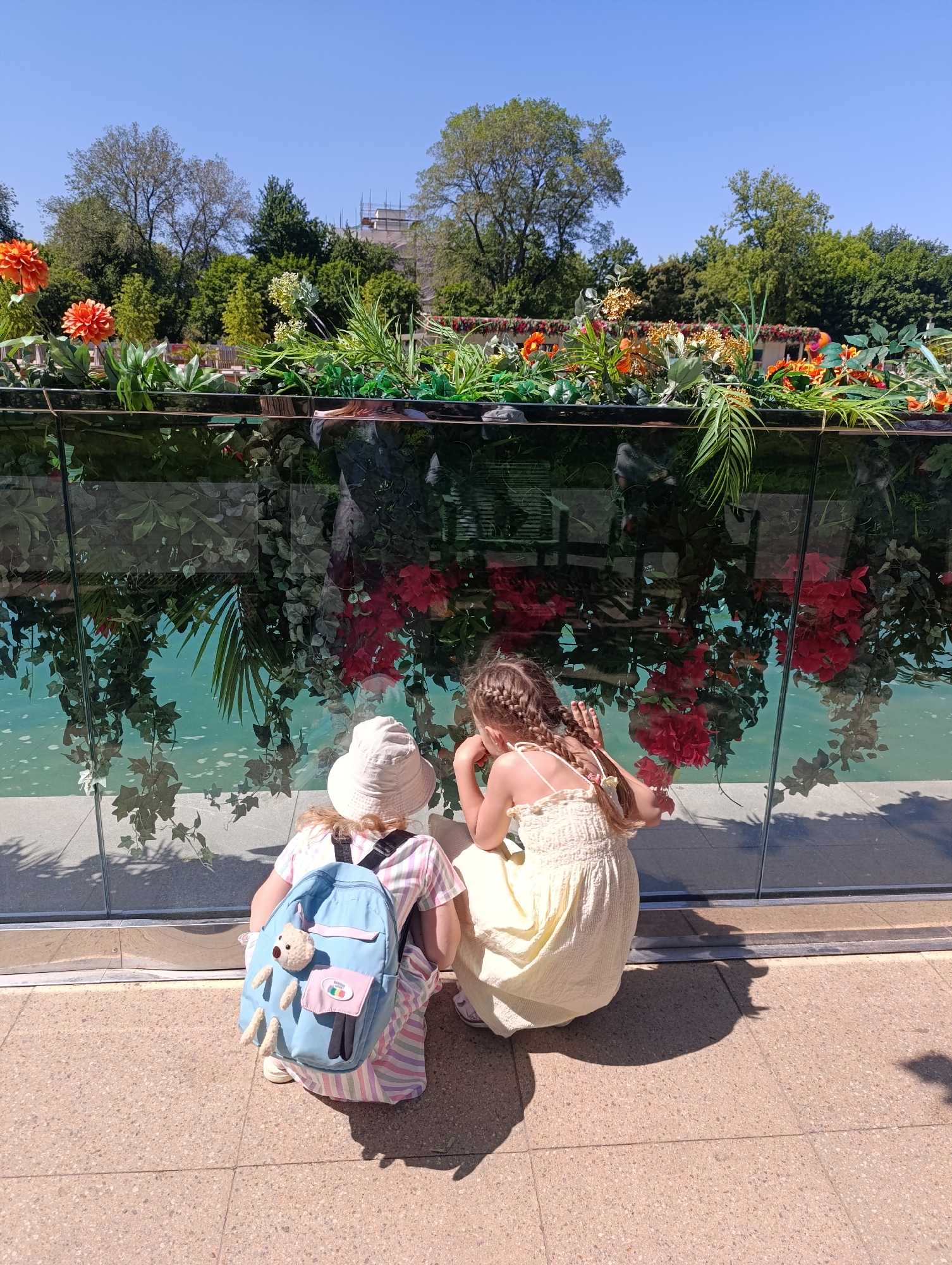 Two young girls are observed from behind, squatting and looking into a transparent glass barrier that fronts a shallow, light blue-green body of water. The scene is outdoors under a clear blue sky, indicating bright daylight.
The girl on the left wears a light-colored bucket hat, a horizontally striped dress in shades of pink, white, and light blue, and a small, light blue backpack adorned with a light brown teddy bear charm and a small pink and white patch. Her hands are not clearly visible.
The girl on the right has her light brown hair styled in two long braids. She wears a light yellow, smocked sundress with thin straps. Her right hand is near her chin, while her left hand rests against the glass barrier.
The glass barrier reflects the sky and various plants positioned above it, creating a visual effect where red, green, and orange foliage appears inverted within the water. Above the barrier, a planter contains lush green plants and vibrant orange flowers, possibly dahlias, along with other floral arrangements.
The background features a dense collection of green trees and, in the upper right, a distant building with visible scaffolding, indicating construction or renovation. The ground beneath the girls is paved with light-colored, speckled square tiles, displaying strong shadows consistent with direct sunlight. The location is an outdoor public space, likely a park or garden within an urban environment.