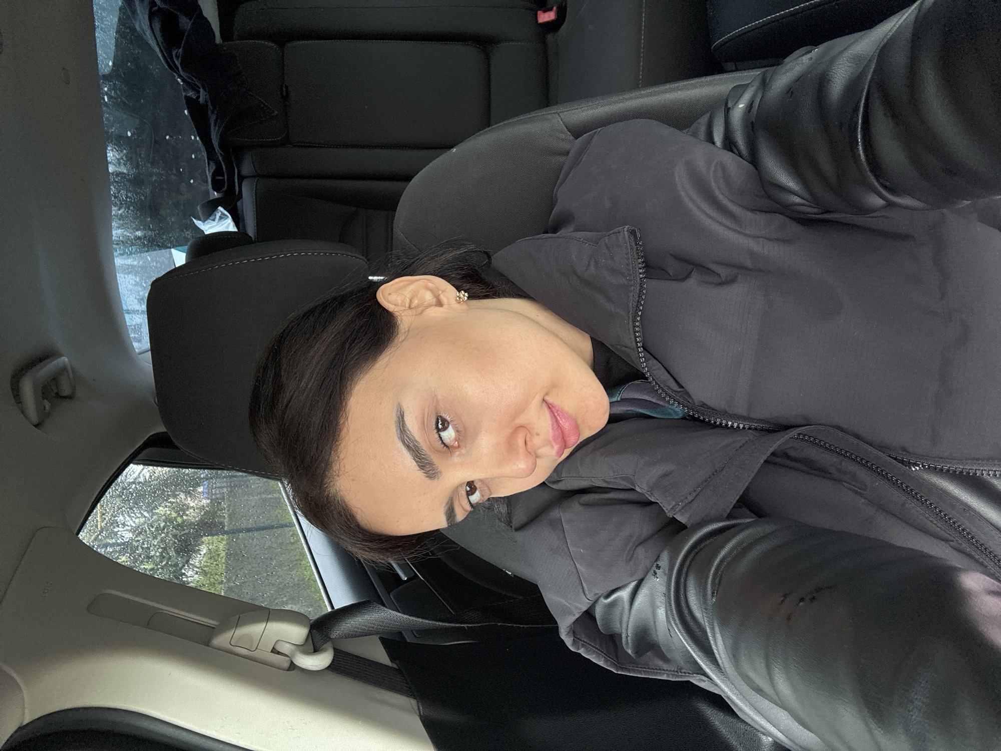 The image depicts a woman inside a car, possibly a passenger in the back seat. She is wearing a black jacket, and the car's interior is primarily black. The view out the window suggests an urban environment, possibly the city of Kartubani, Georgia. The weather appears to be overcast.