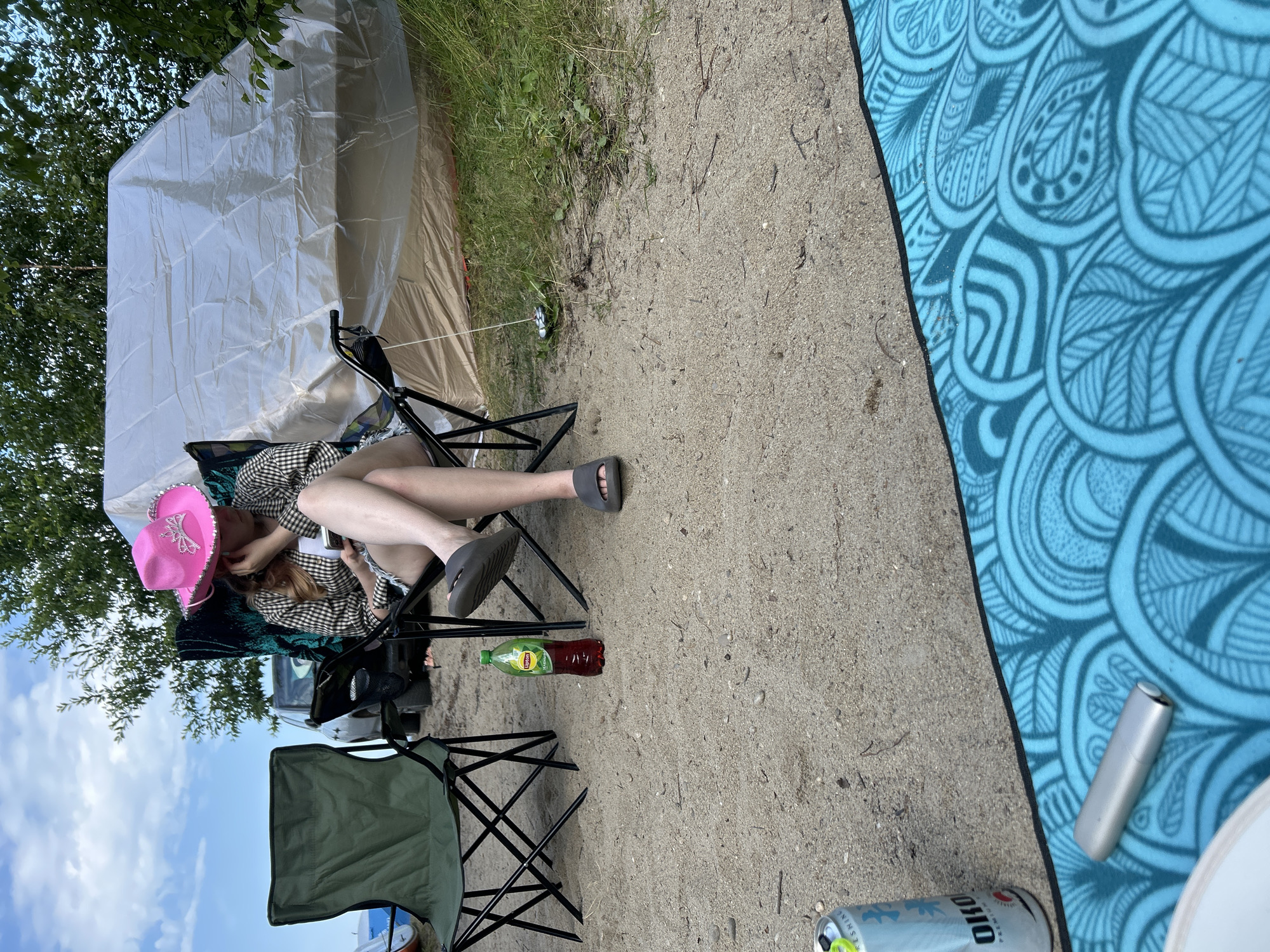 A person wearing a pink cowboy hat sits in a camping chair on a sandy area, looking at a phone. A bottle of Lipton iced tea is on the ground next to them. In the background, a large white tent is set up, and trees are visible. A green camping chair is also present. A can and a silver object are on a blue patterned mat in the foreground. The scene appears to be a campsite.